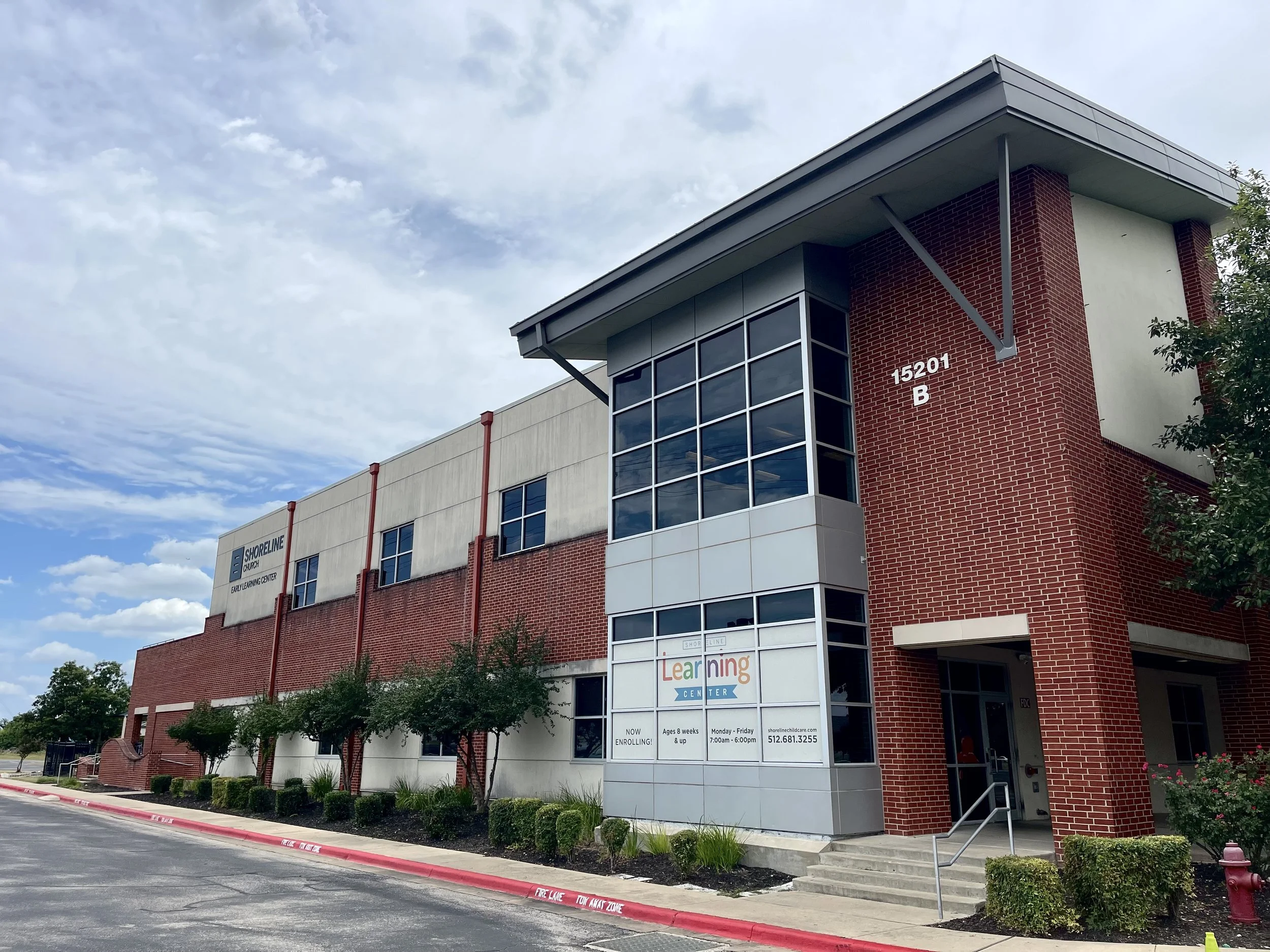 Exterior view of a two-story building with red brick and white walls, housing a learning center with a sign and purple letters. The building's address is 15201 B, and there are small trees and bushes along the sidewalk under a partly cloudy sky.