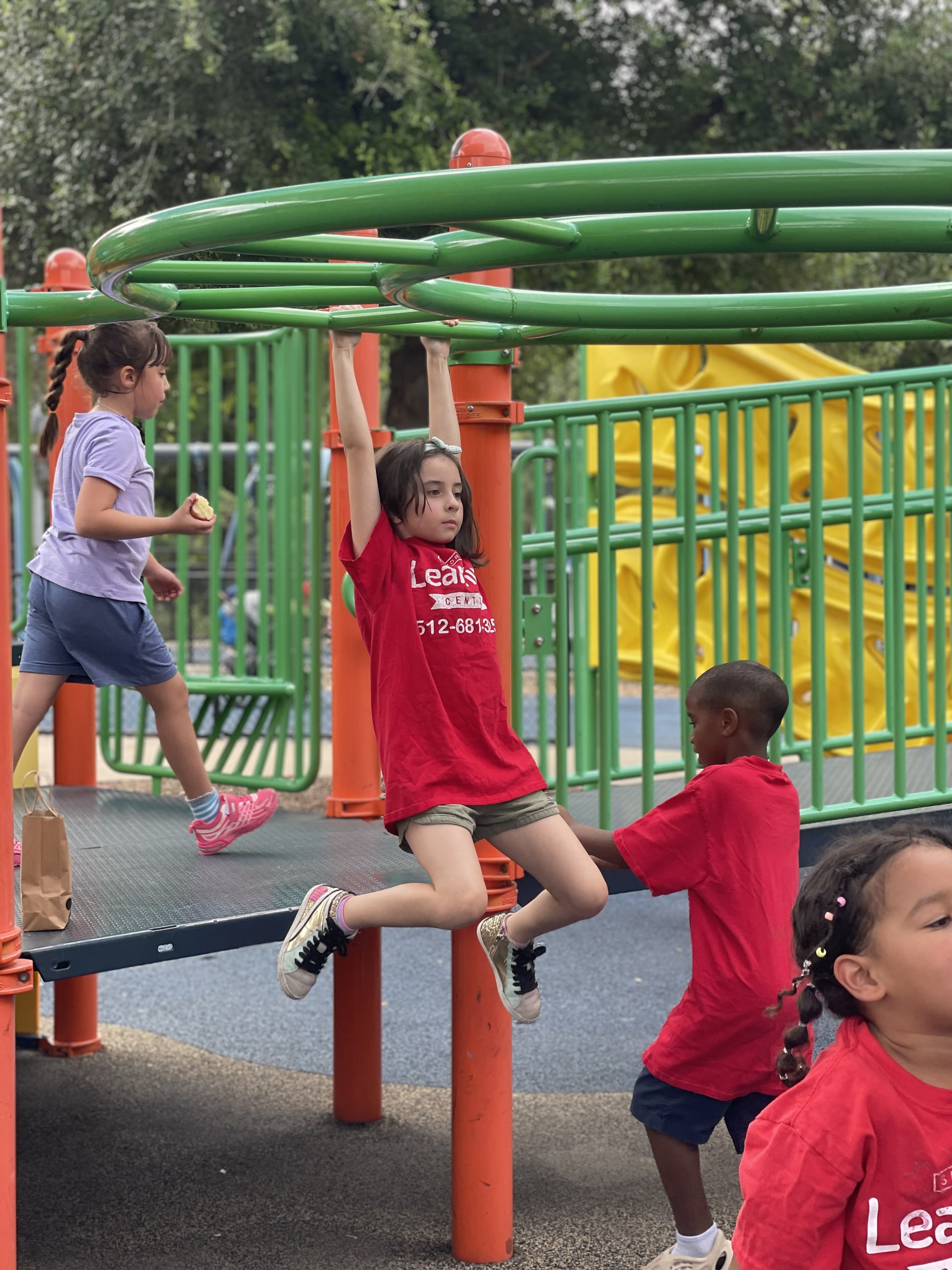 Children playing on a colorful playground with green and yellow equipment, surrounded by trees.