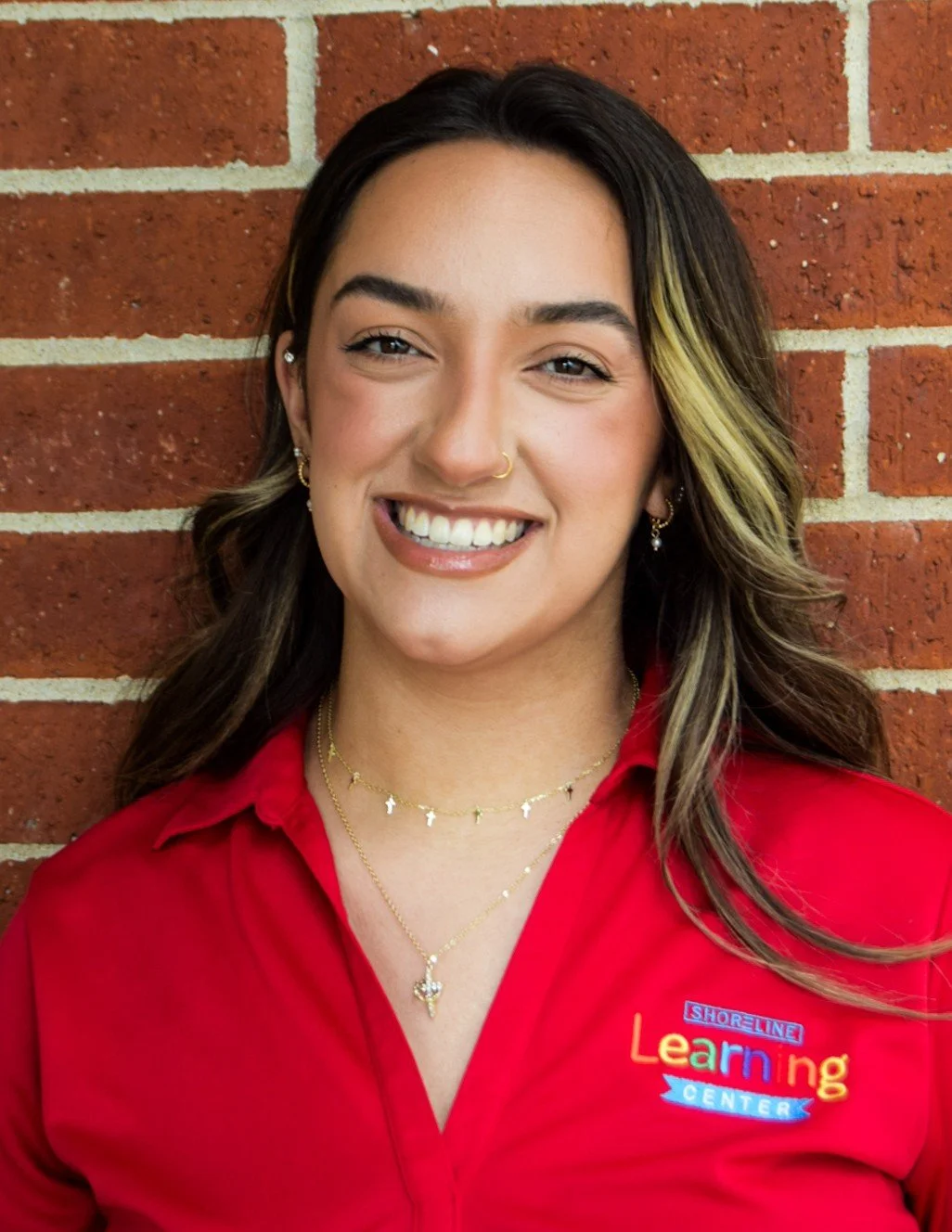 A young woman with dark hair streaked with blonde highlights, smiling, wearing a red shirt with a 'Shoreline Learning Center' embroidery, standing in front of a brick wall.