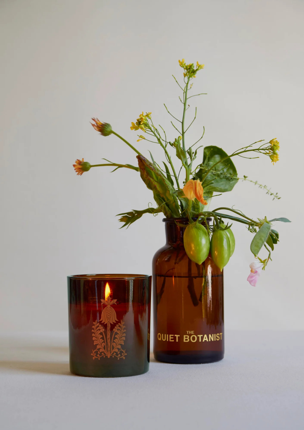 Glowing lit Lit Home Grown Candle glowing warmly beside vase of flowering tomato plants