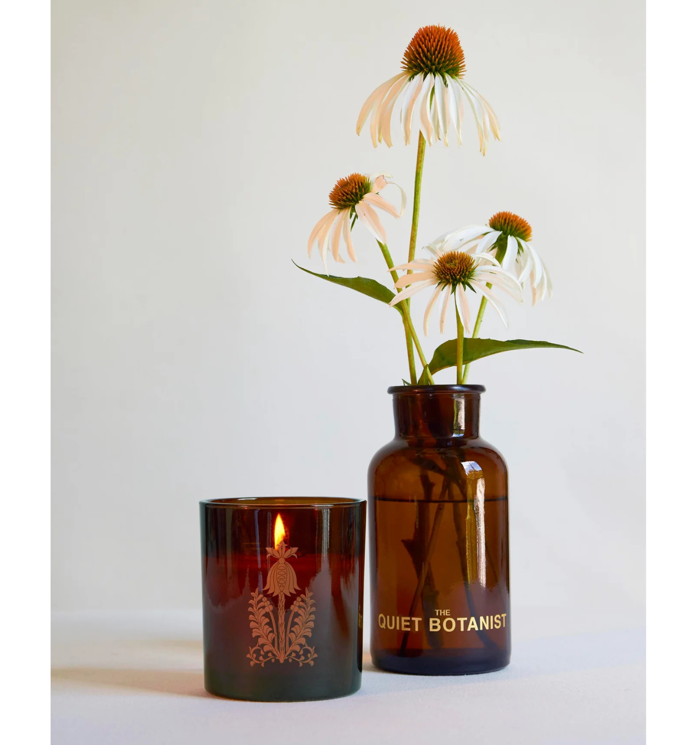 In the Weeds 14 oz lit candle in brown glass next to vase of flowers