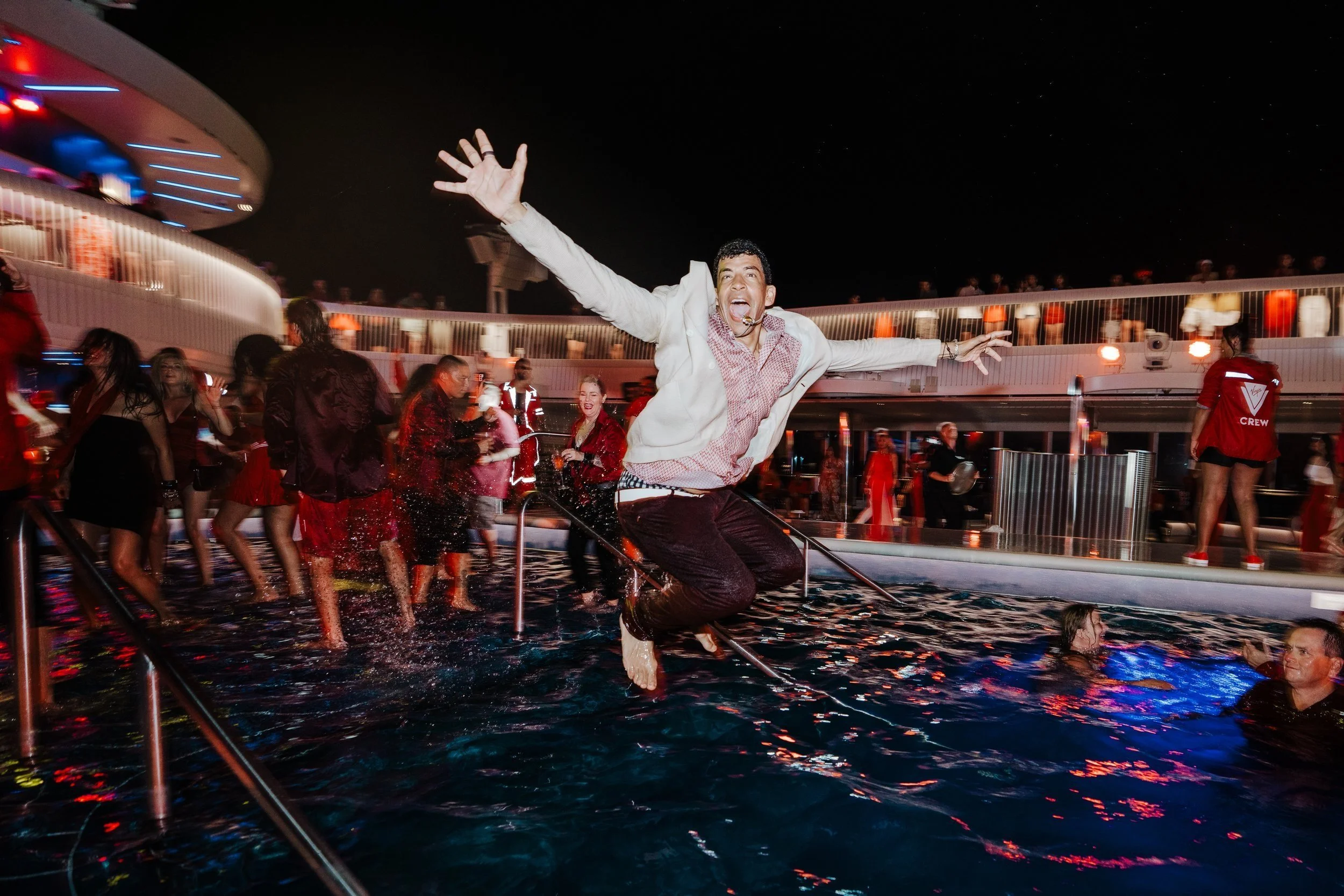 A man jumping into a swimming pool with arms outstretched, smiling, at a nighttime party with many people in red and festive attire, on a cruise ship deck.