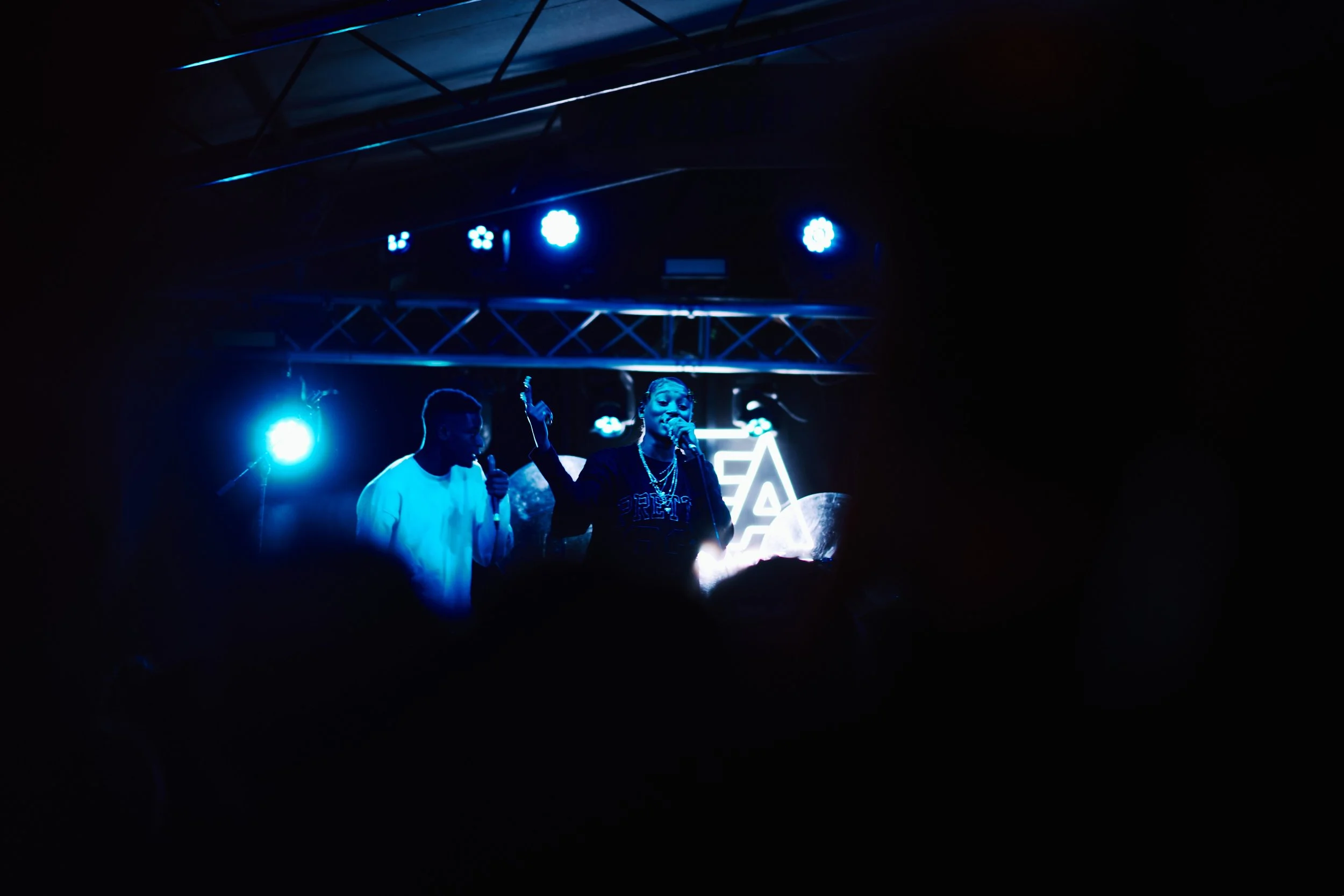 Two performers on stage under blue lighting, one singing into a microphone with a raised hand, the other standing nearby with a microphone, against a backdrop with abstract designs.