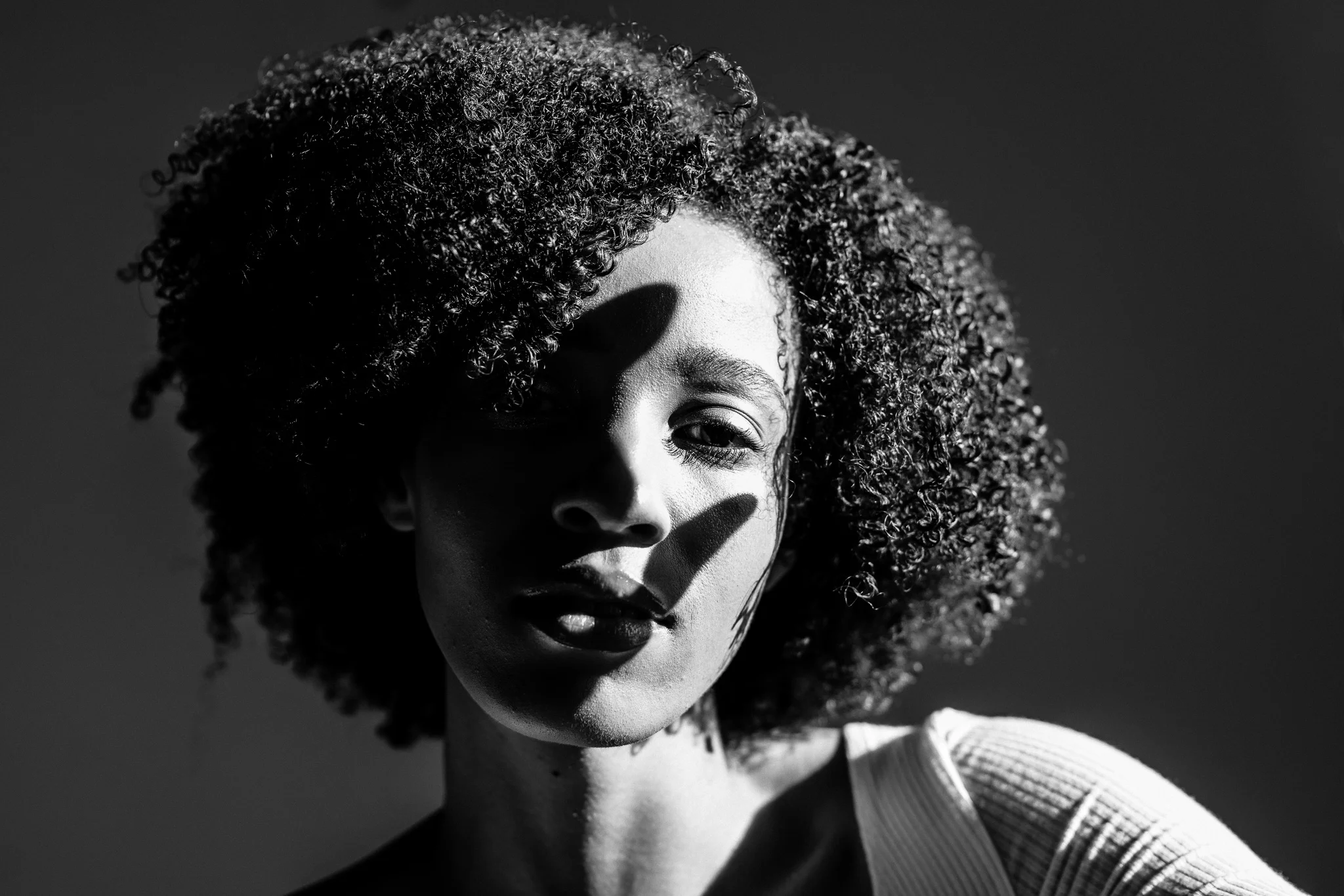 A black and white portrait of a woman with curly hair, partially shadowed, wearing a light-colored top.