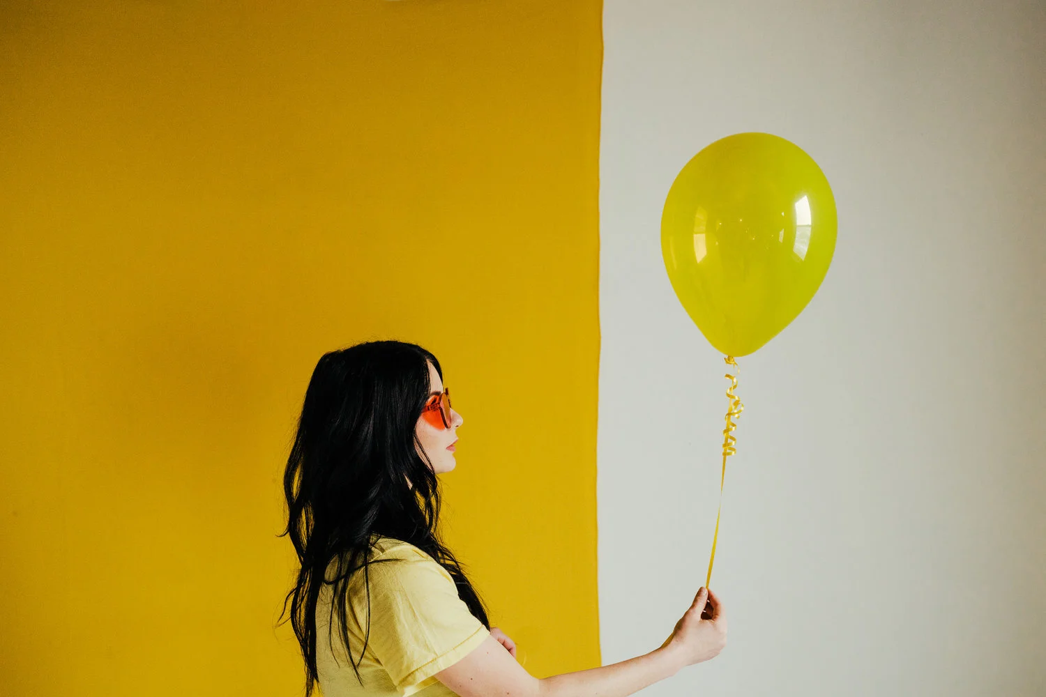 A woman with long black hair wearing orange sunglasses and a yellow shirt holding a yellow balloon with the name 'Milan' written on the string, standing against a yellow and white wall.