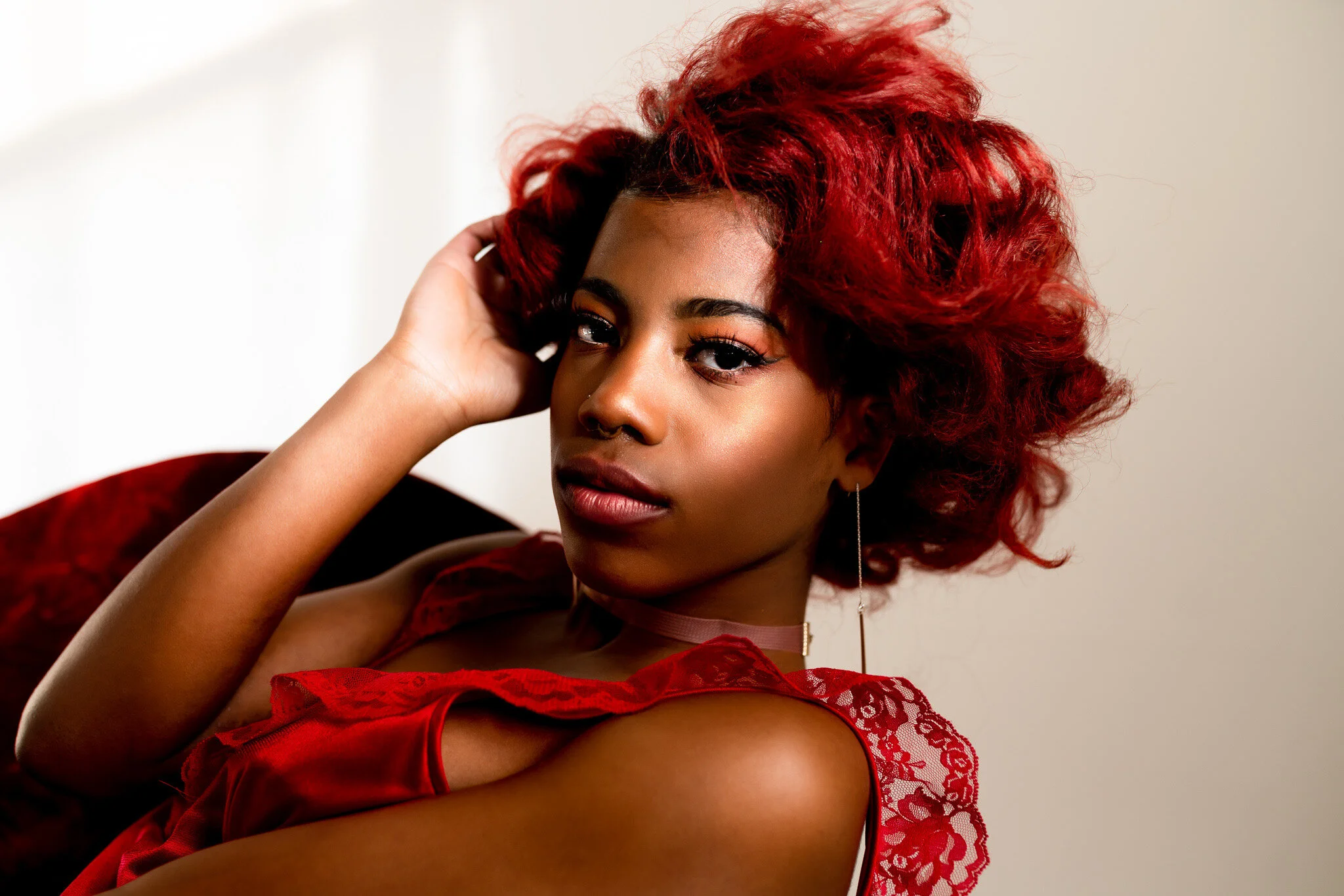 A woman with curly red hair, wearing a red lace dress and long earrings, is posing indoors with her hand resting on her head, looking directly at the camera.