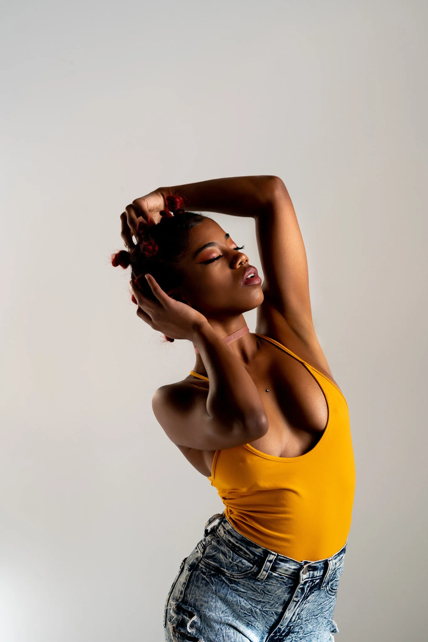 A woman with dark skin and red bantu knots hairstyle, wearing a yellow tank top and distressed denim shorts, posing with her eyes closed and hands on her head against a plain background.