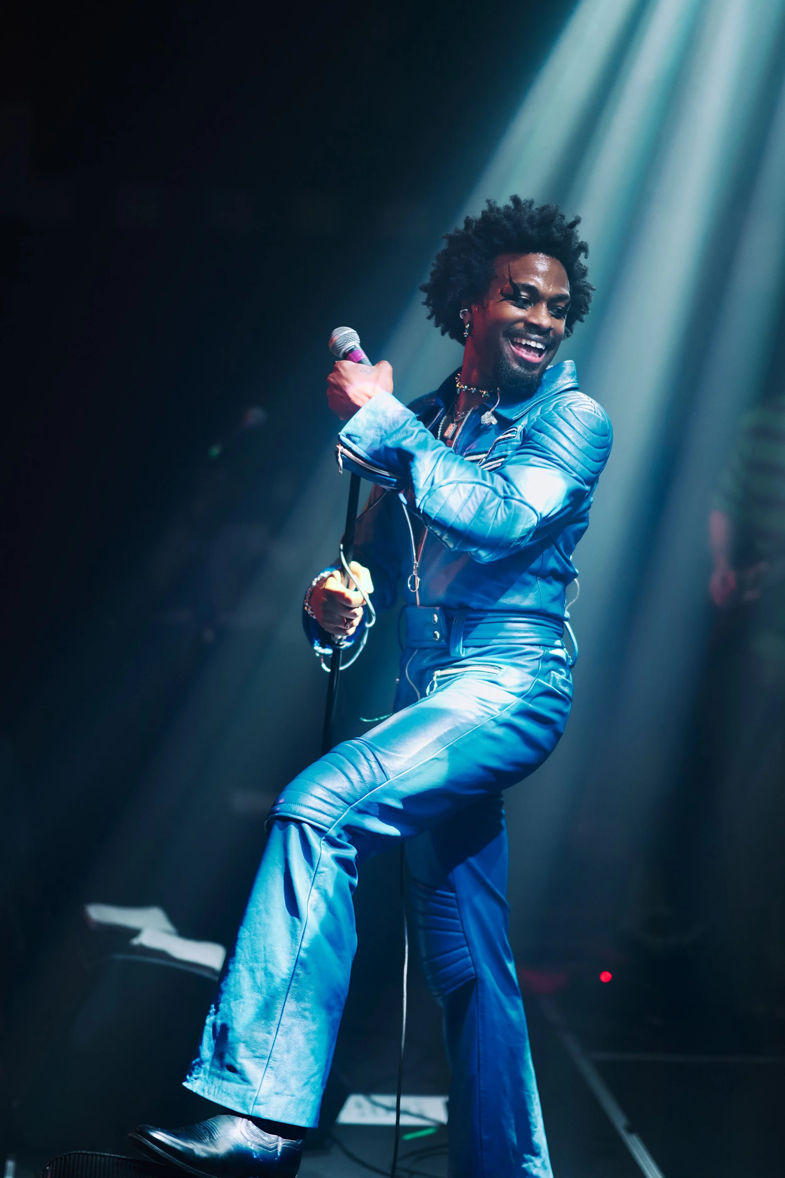A man with curly hair, dressed in a shiny blue leather outfit, is singing or speaking into a microphone on stage, illuminated by dramatic stage lighting.