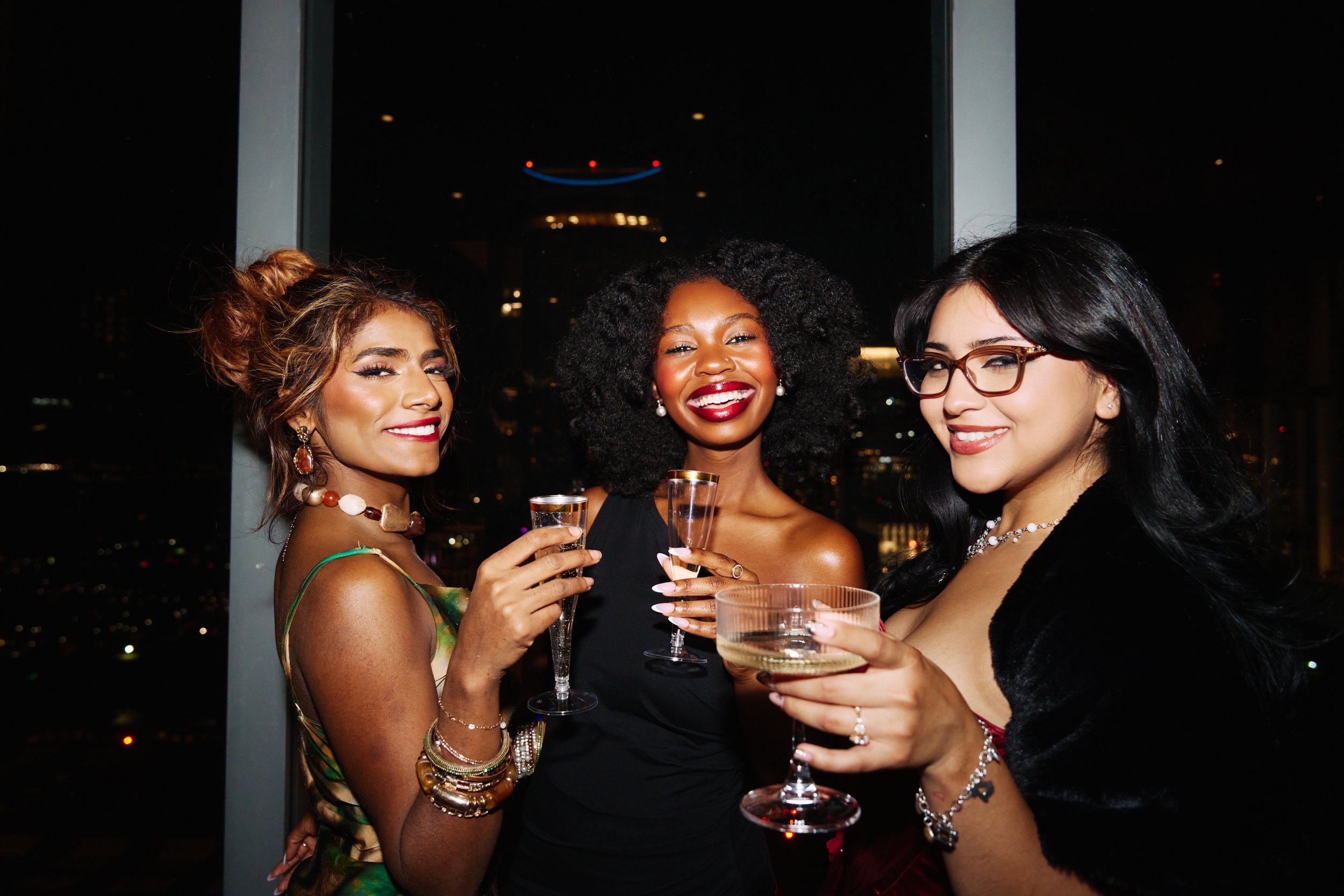 Three women in elegant dresses holding glasses of champagne at a nighttime social event, smiling and enjoying themselves in Austin, Texas