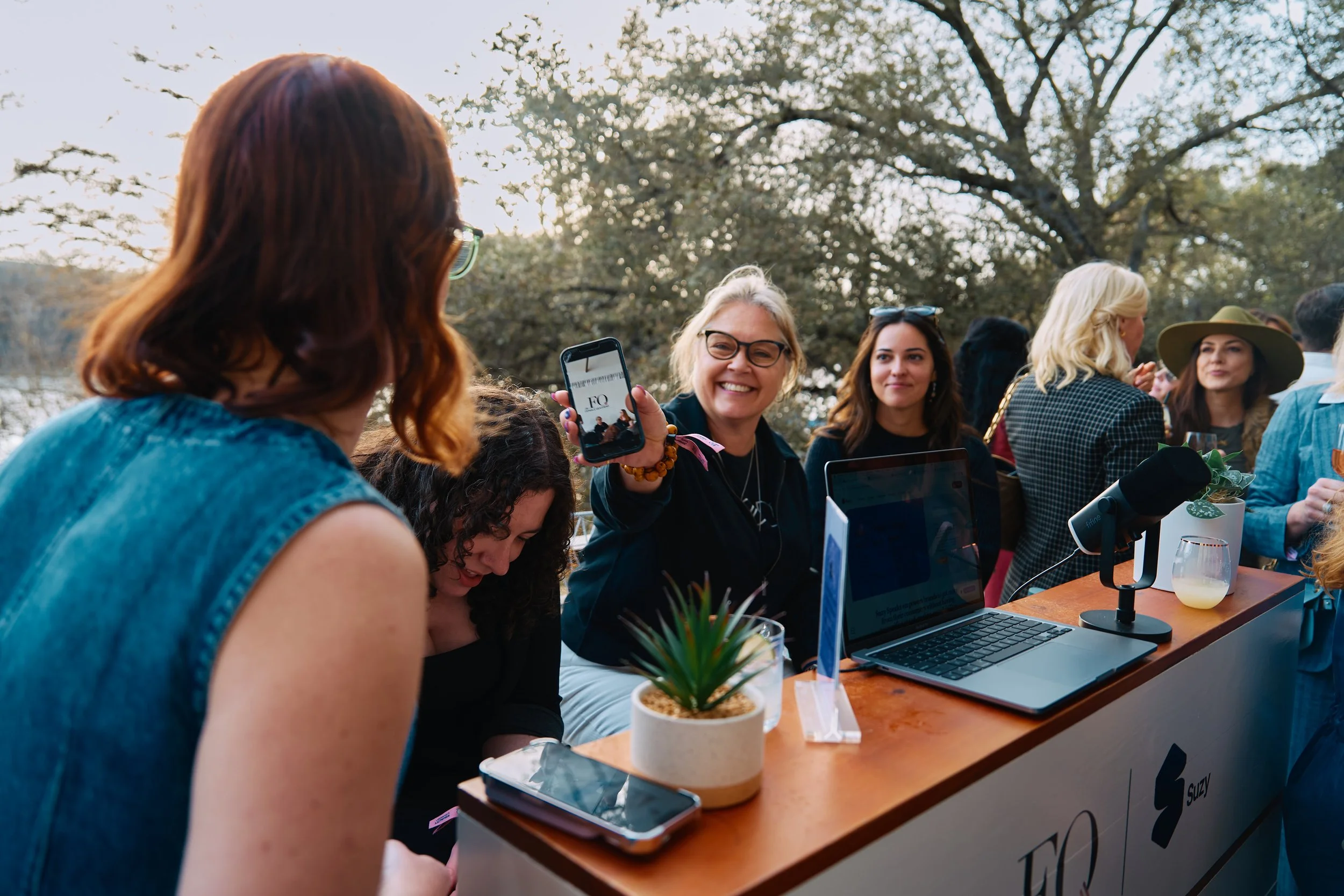 Group of women at an outdoor event, one woman showing her phone with a cheerful smile, others gathered around a table with laptops, drinks, and a potted plant, trees in the background during SXSW Event in Austin, TX.
