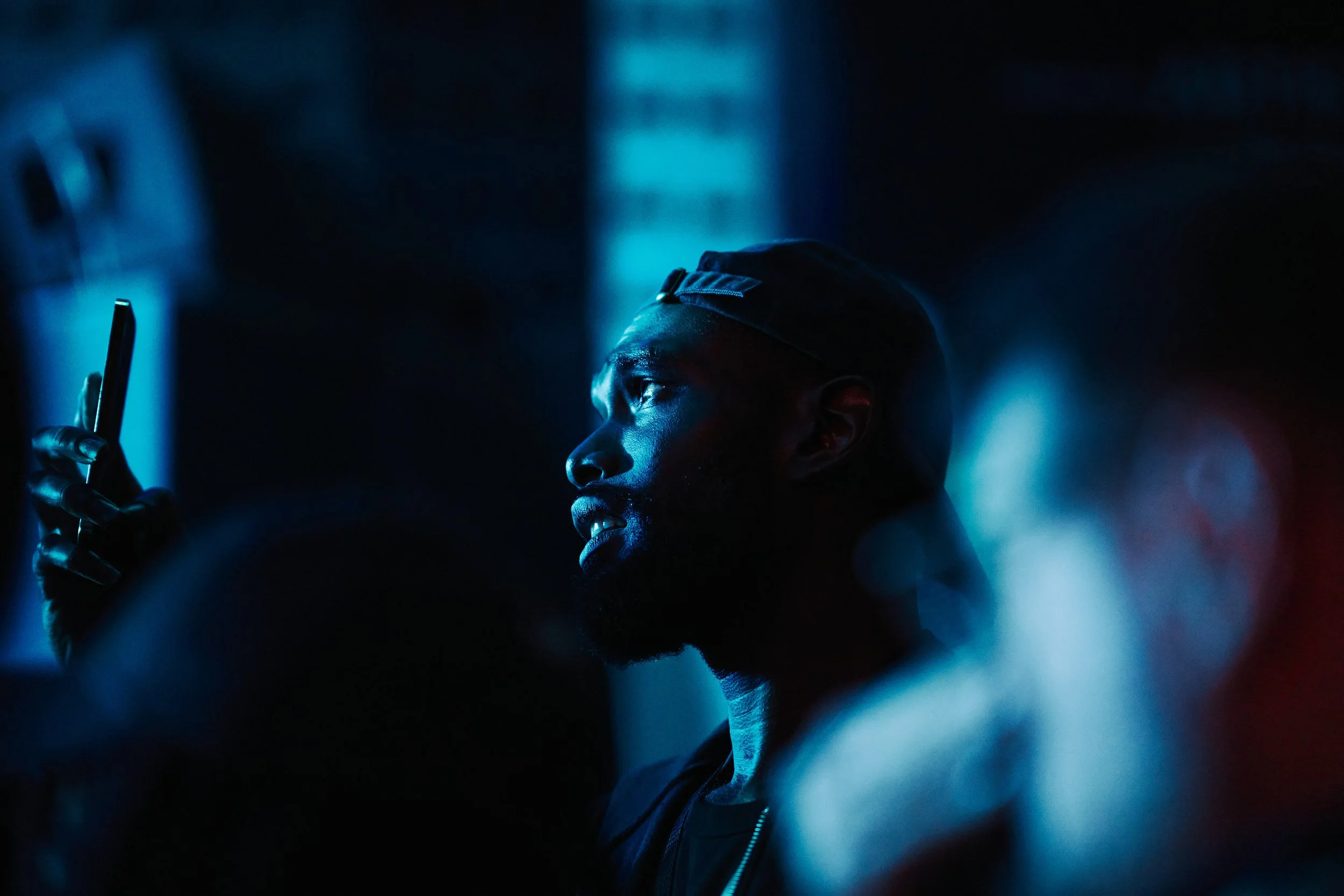 A man wearing a dark hat and jacket is illuminated by blue and red neon lights, looking at his smartphone in a dark environment.