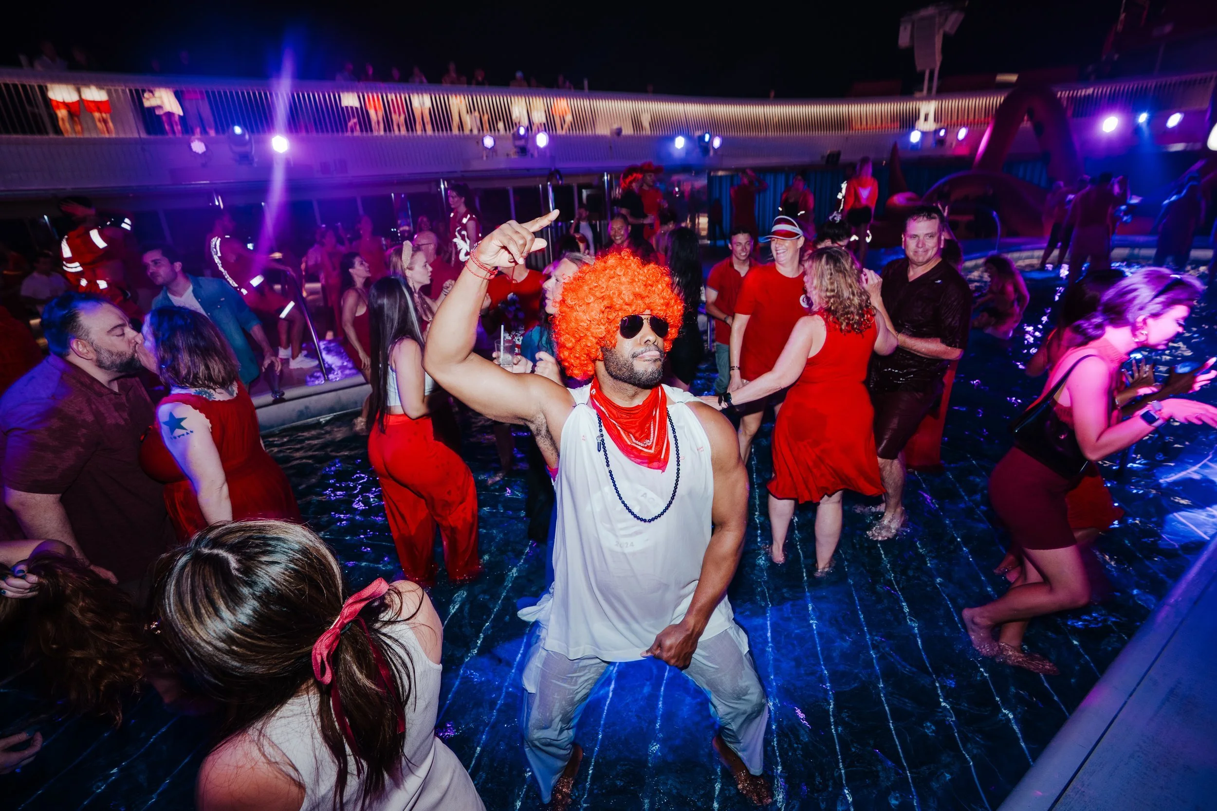 A man dressed as a clown with an orange curly wig, sunglasses, white sleeveless shirt, and red bandana dancing in a lively pool party with other guests, some dressed in red, under colorful lights.
