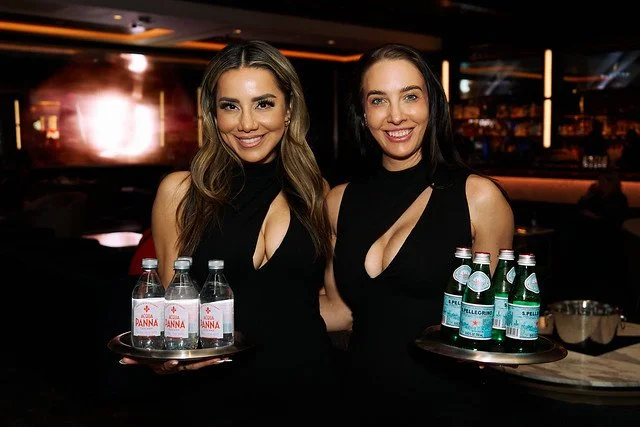 Two women wearing black dresses holding trays of bottled water in a dimly lit restaurant or bar.