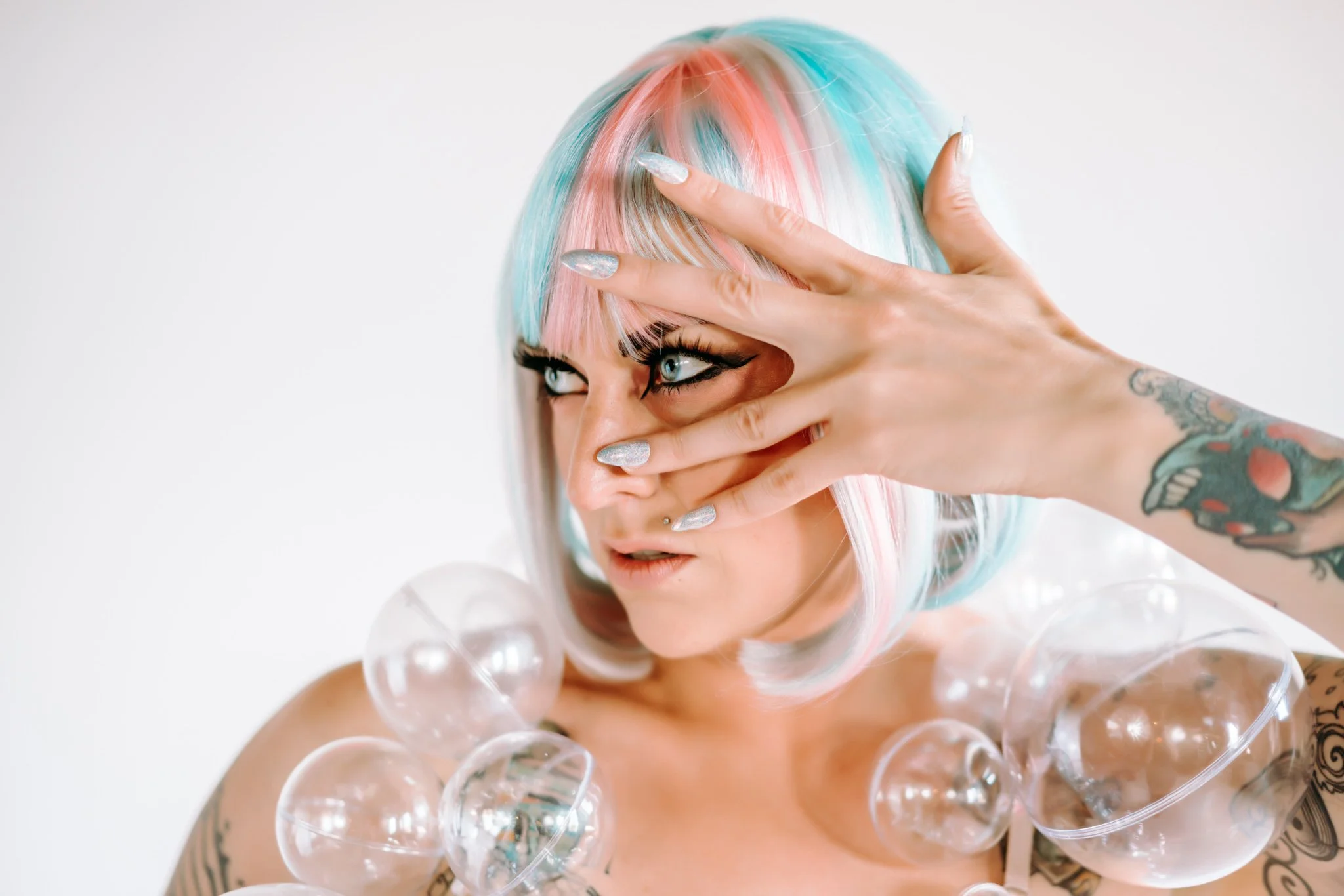 A woman with pastel multicolored hair and tattoos, looking to her left with an intense expression, holding her hand in front of her face, surrounded by transparent plastic bubbles.