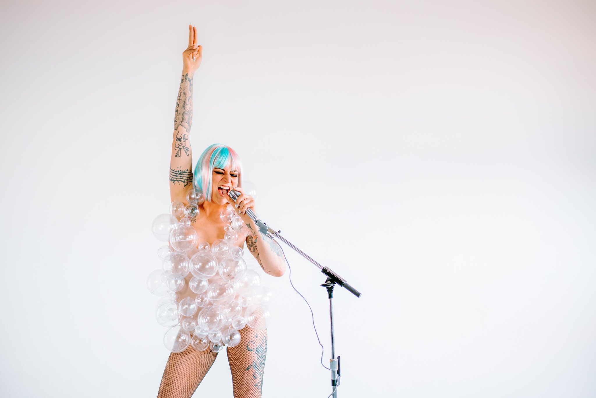 A woman with colorful hair and tattoos singing into a vintage microphone, wearing a costume made of clear plastic bubbles, in front of a plain white background.