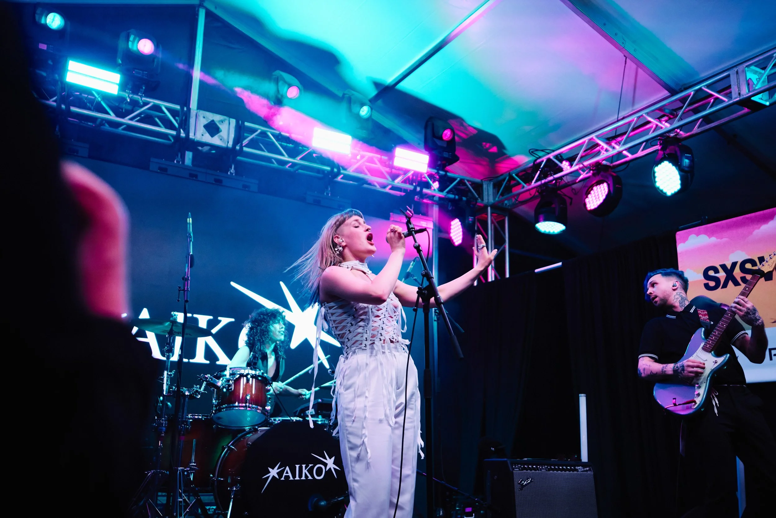 A live musical performance on stage featuring a woman singing into a microphone with her eyes closed, accompanied by a drummer and a guitarist. Stage lights illuminate the scene with pink, purple, and blue hues, with a black backdrop and the SXSW logo visible.
