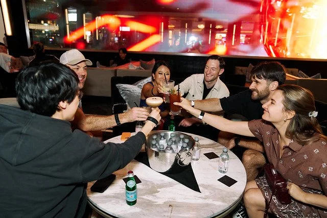 Group of six young adults sitting around a table, enjoying drinks and toasting in a lively bar or lounge.