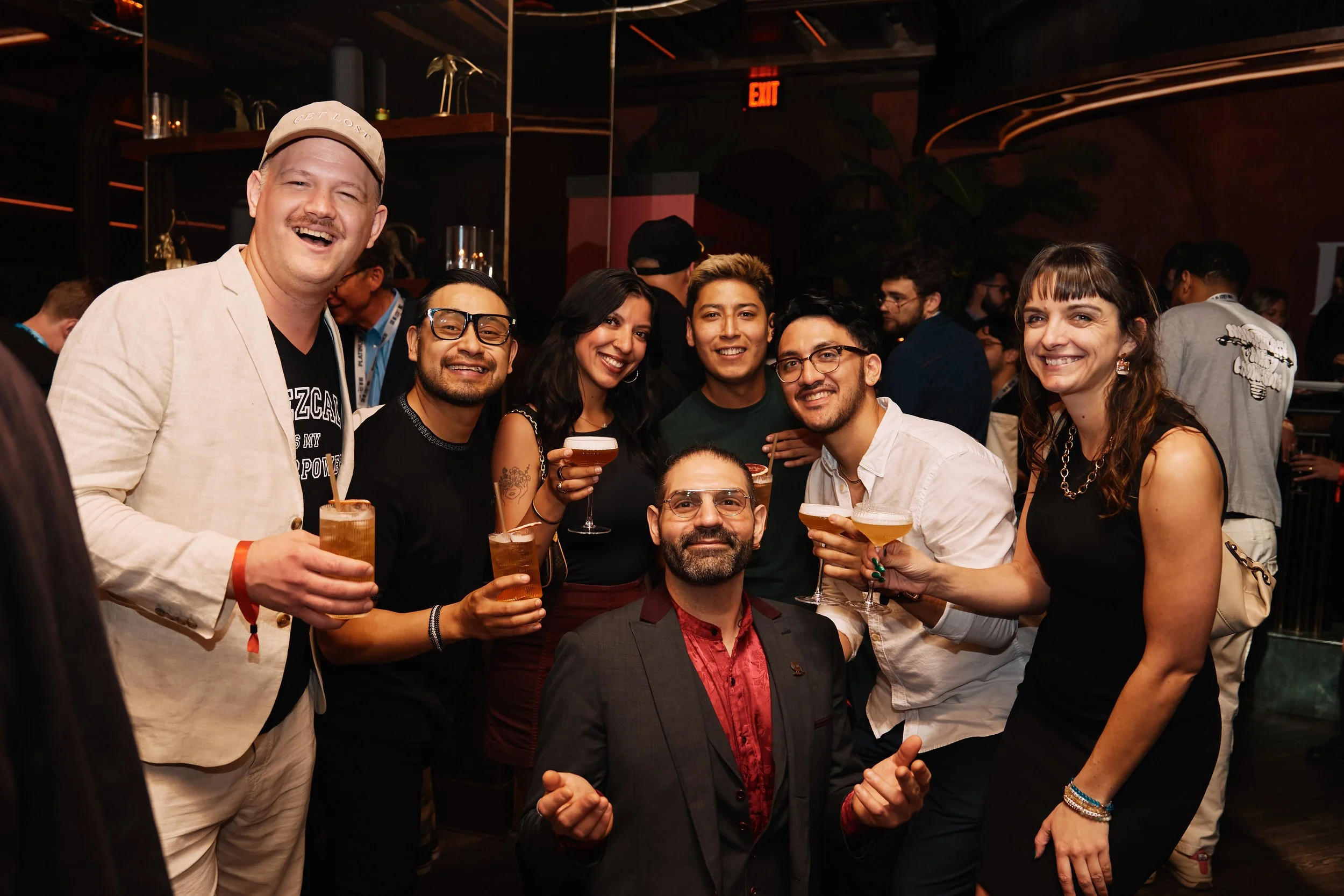 Group of seven diverse adults smiling and holding cocktails at a party or bar, with a dark indoor background. during SXSW Event in Austin, TX