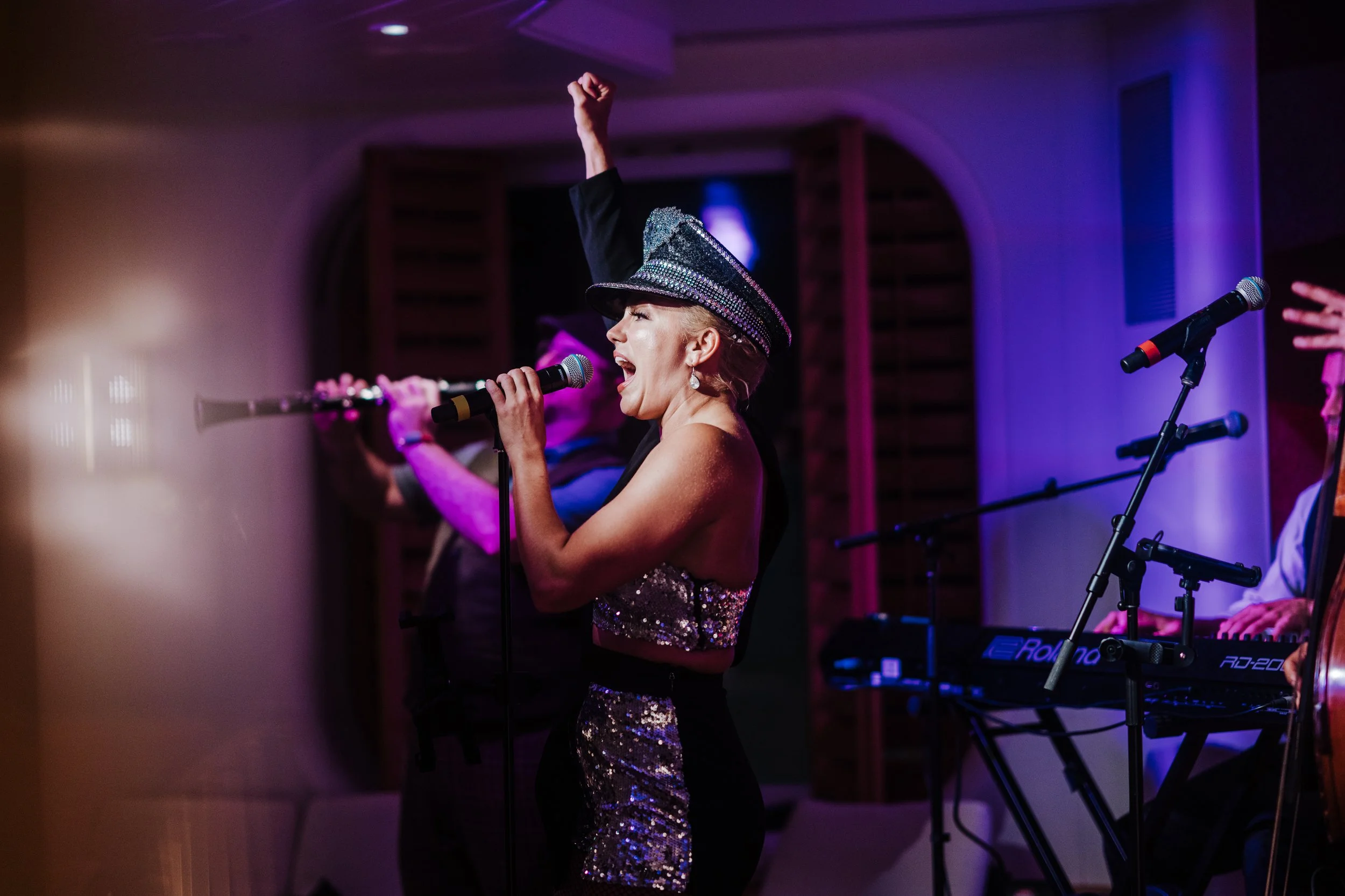 A woman singing into a microphone at a live music event, wearing a sparkly outfit and a sequined hat, with a keyboard and musicians in the background.
