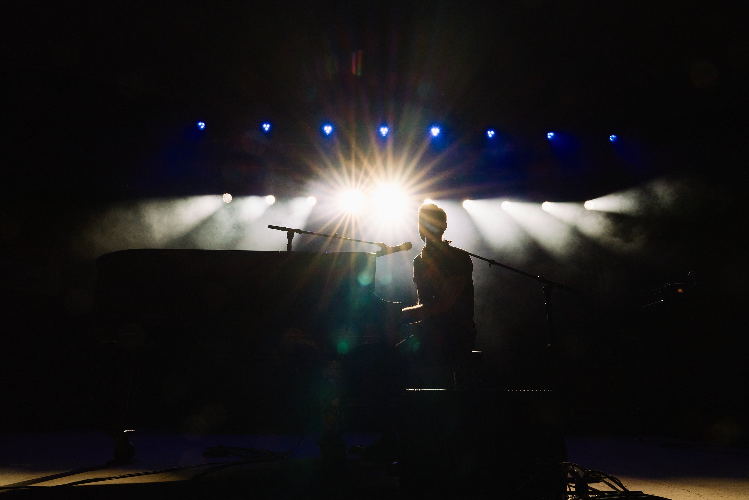 Silhouette of a performer at a piano with bright stage lights shining behind, creating a starburst effect, in a dark concert setting.