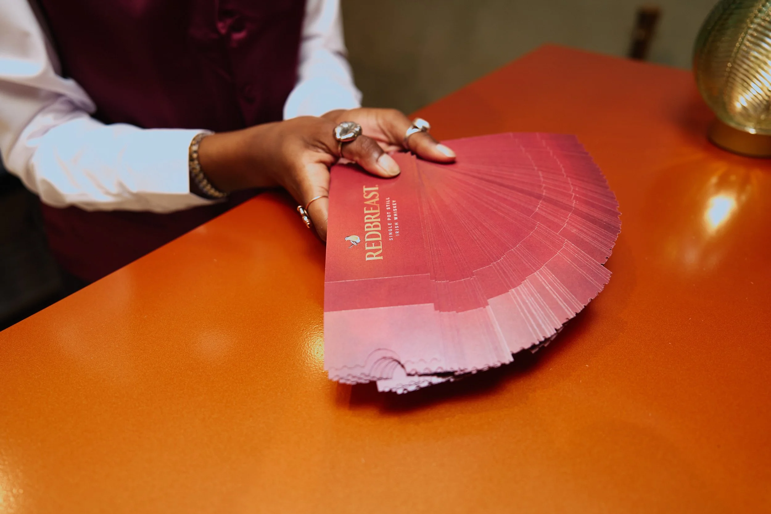 A person holding a pink paper fan with the text 'REDBREAST' on it, sitting at an orange table with a gold decorative object to the right during SXSW Event in Austin, TX