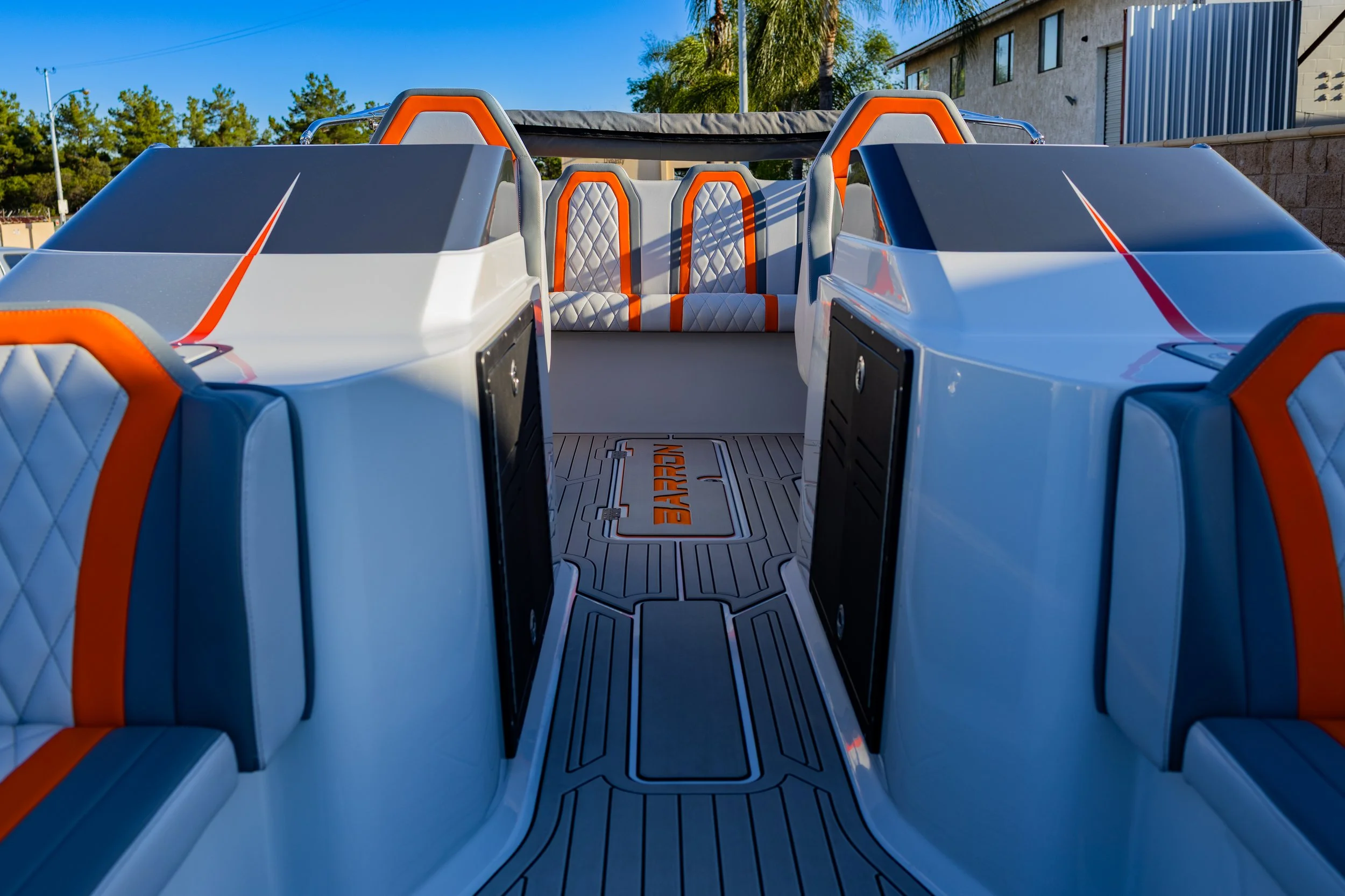 Interior of a futuristic speedboat with white and orange seats and a wooden floor, with trees and a building in the background.