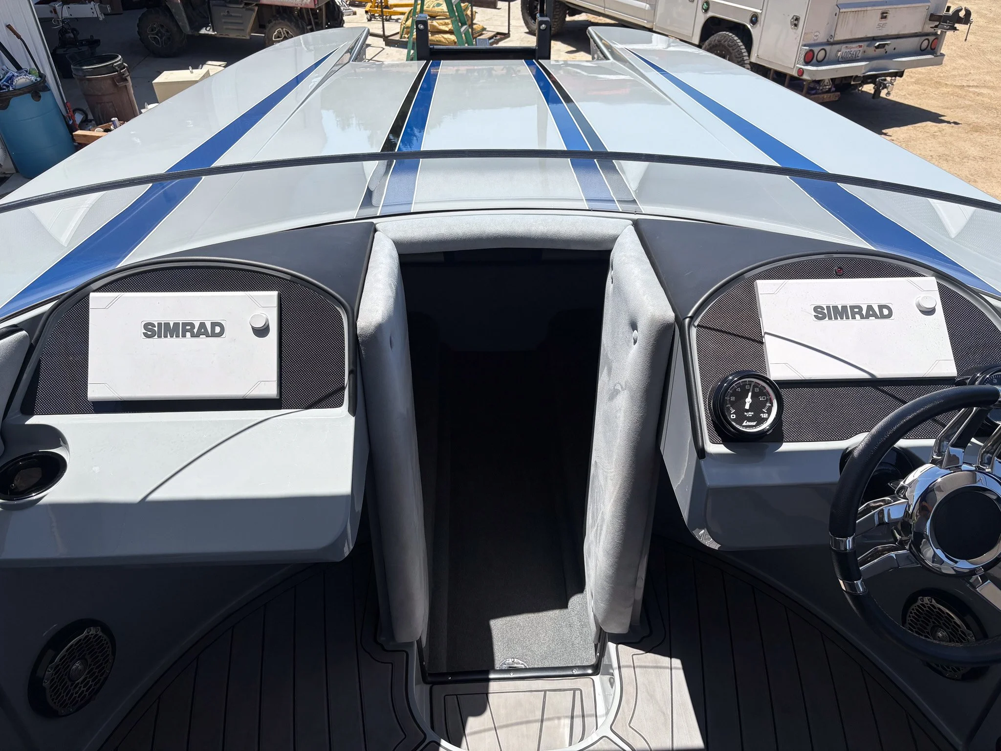 The image shows the dashboard and interior of a boat with Simrad electronic systems and steering wheel, with various equipment and vehicles visible outside the boat.