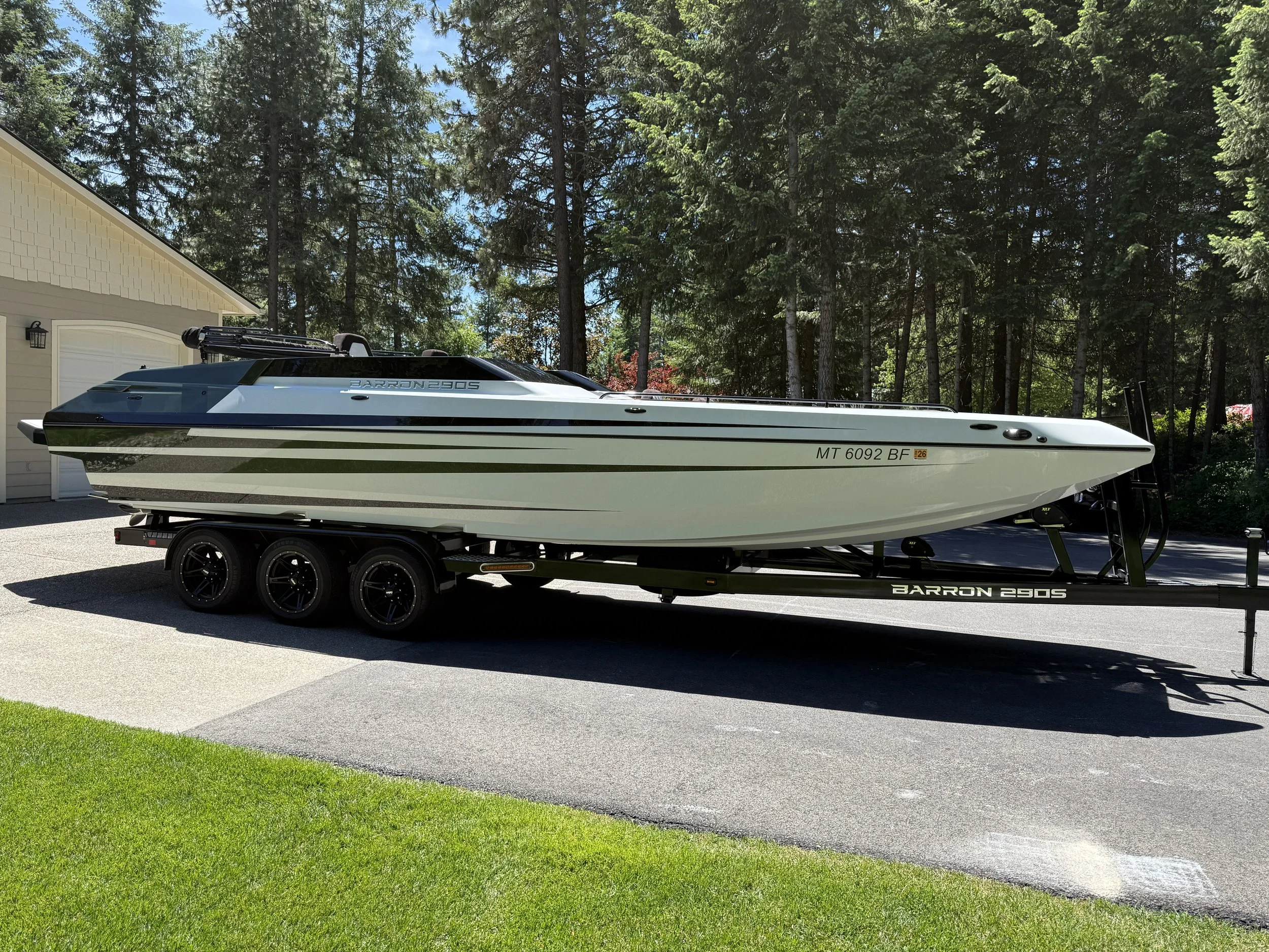 A speedboat on a trailer parked in a driveway with a house and a dense forest of tall green trees in the background.