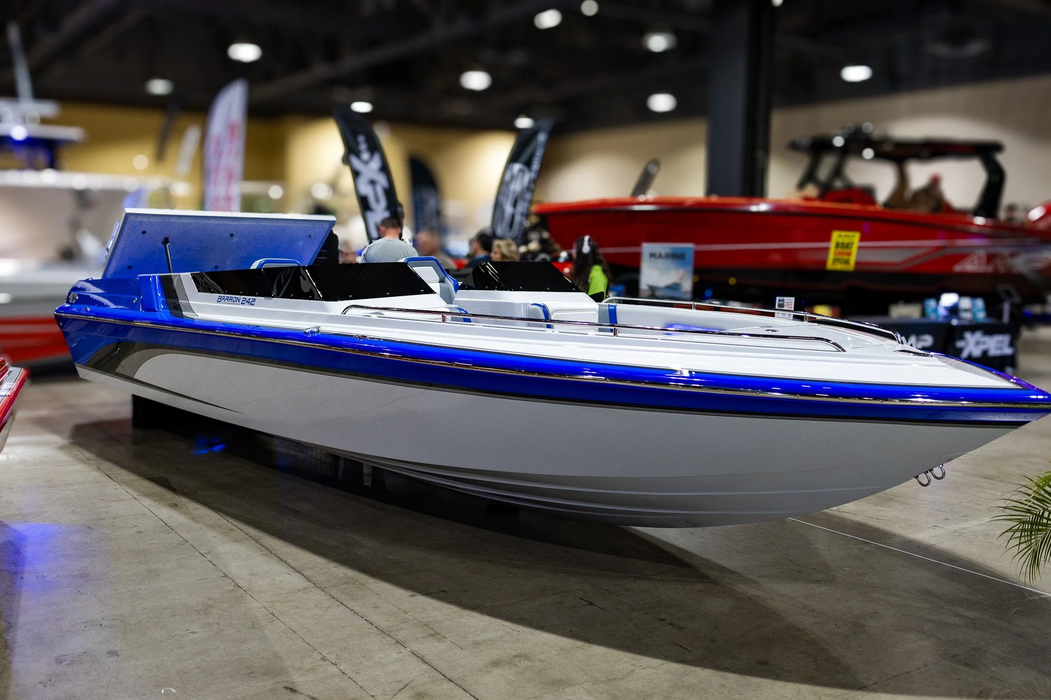 A sleek blue and white speedboat displayed at an indoor boat show with other boats and people in the background.