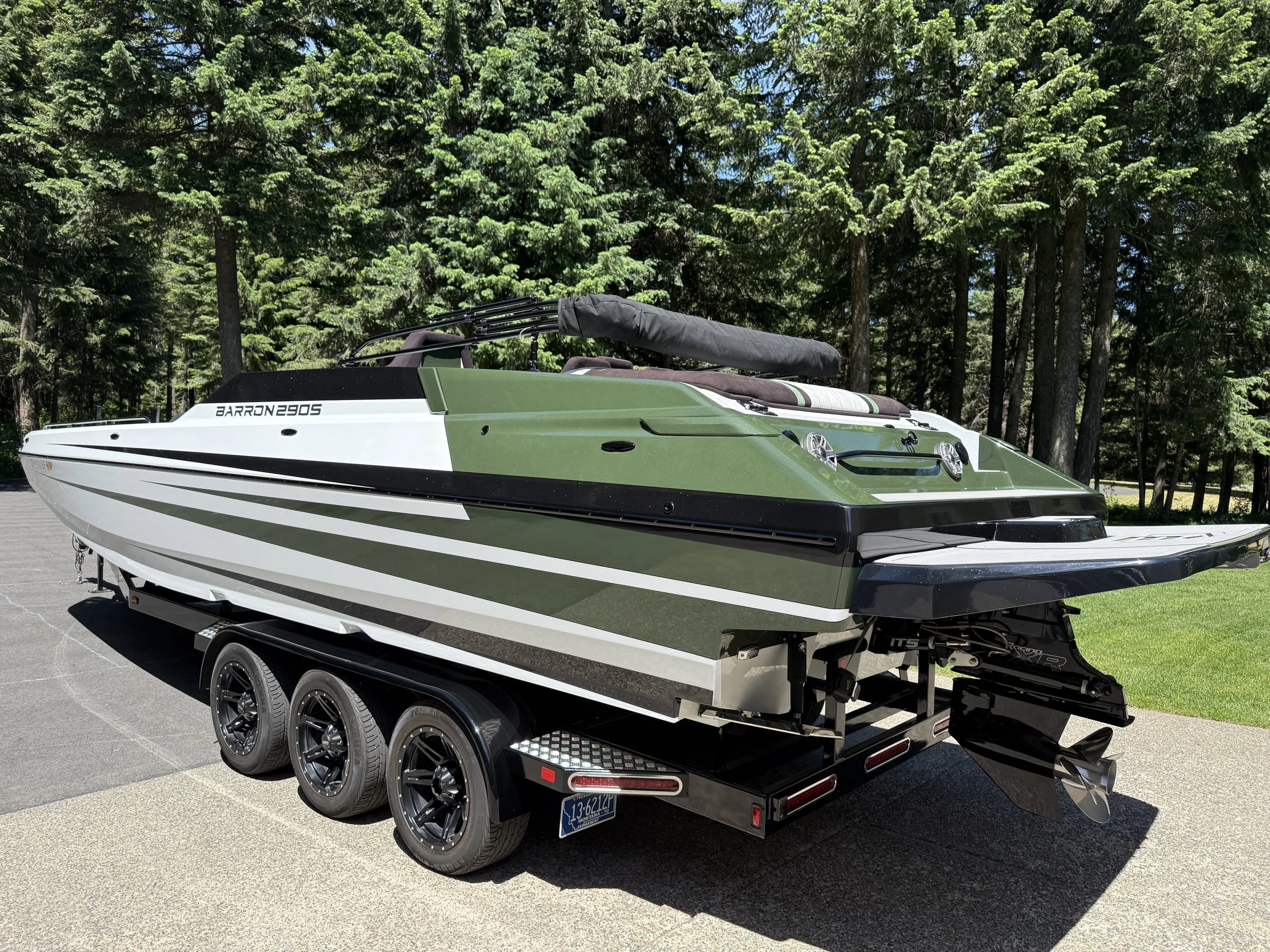 A green and white speedboat on a trailer parked on a paved surface with a background of tall green trees.