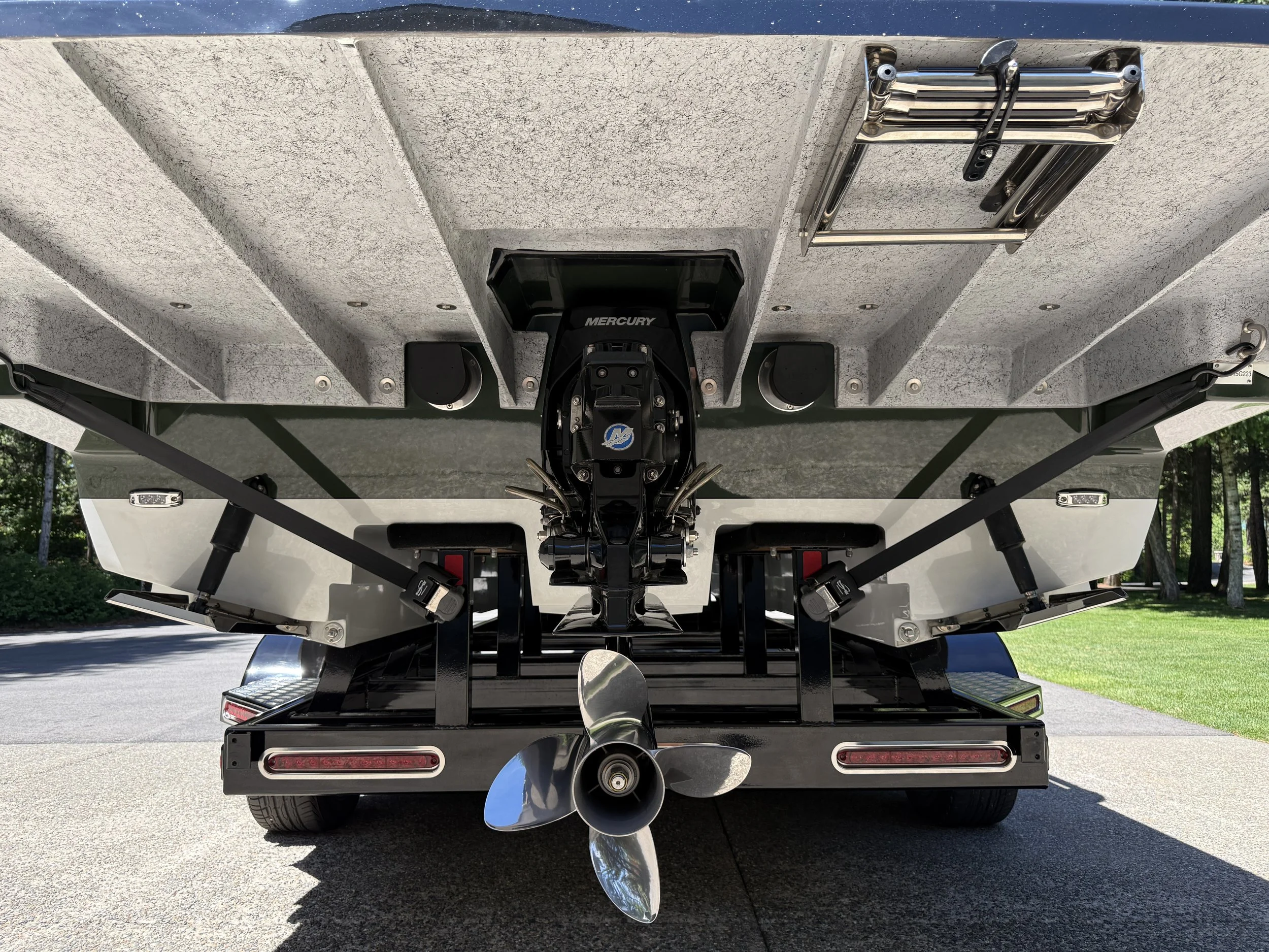 Rear view of a boat trailer with a Mercury outboard motor, a propeller, and boat accessories mounted on the underside of a boat, parked on a paved surface.