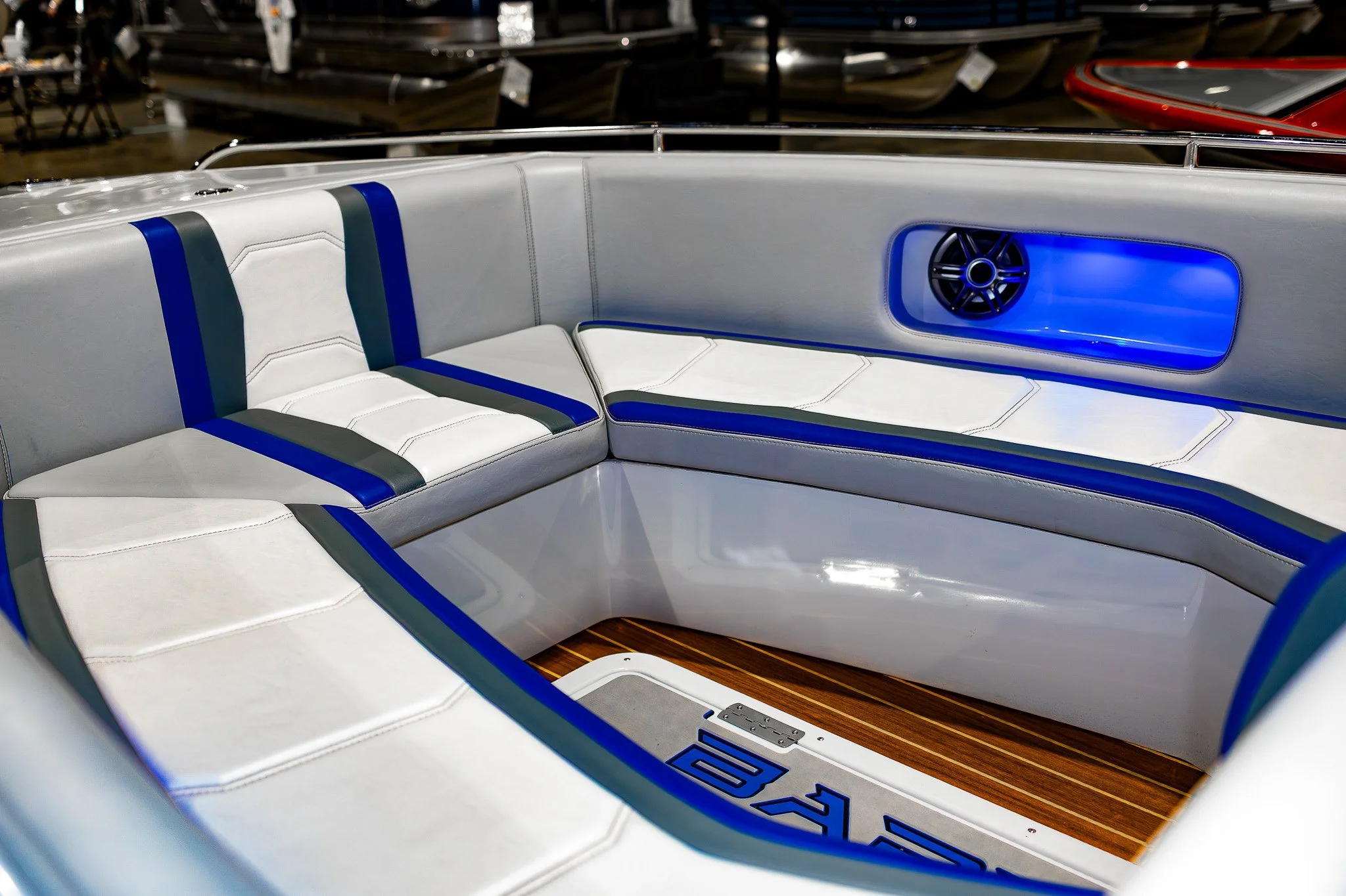 Close-up of the seating area on a boat, with white and blue cushioned seats, a small speaker with blue accent lighting, and a teak floor section.