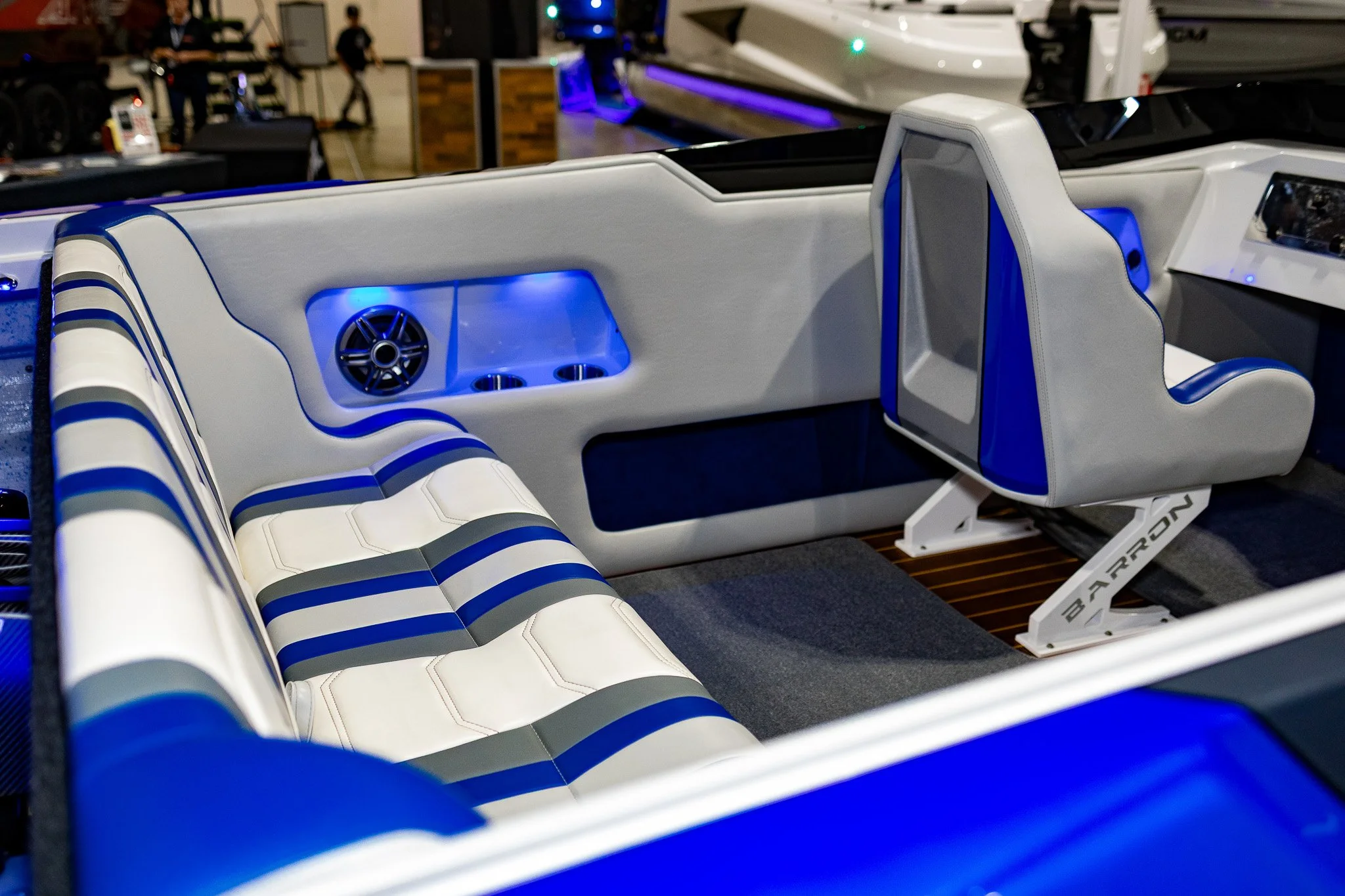 Interior of a luxury boat with white and blue cushioned seating, a seat with a high backrest, built-in speakers, and cup holders, illuminated by blue lighting.
