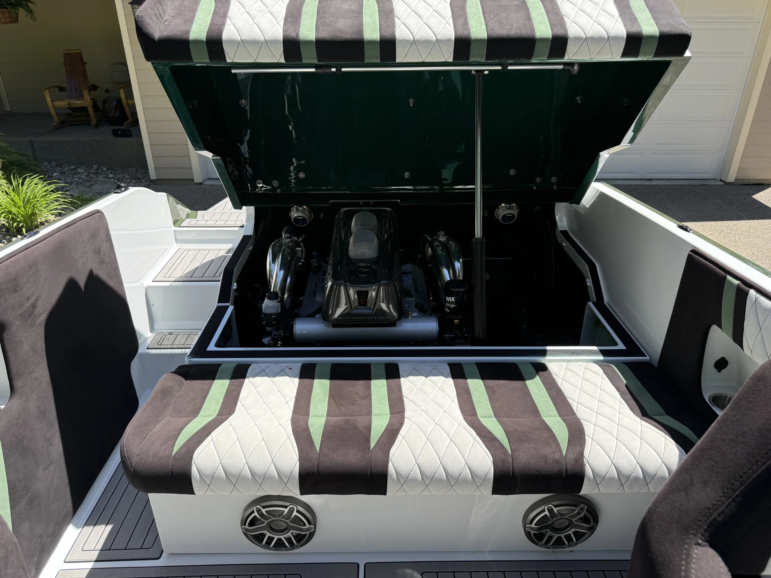 Front view of a golf cart with a black hood lifted, showing the engine inside. The golf cart has cushioned seats with green, brown, and white striped covers, and speakers below the seats. It is parked outside near a house with a garage, on a concrete