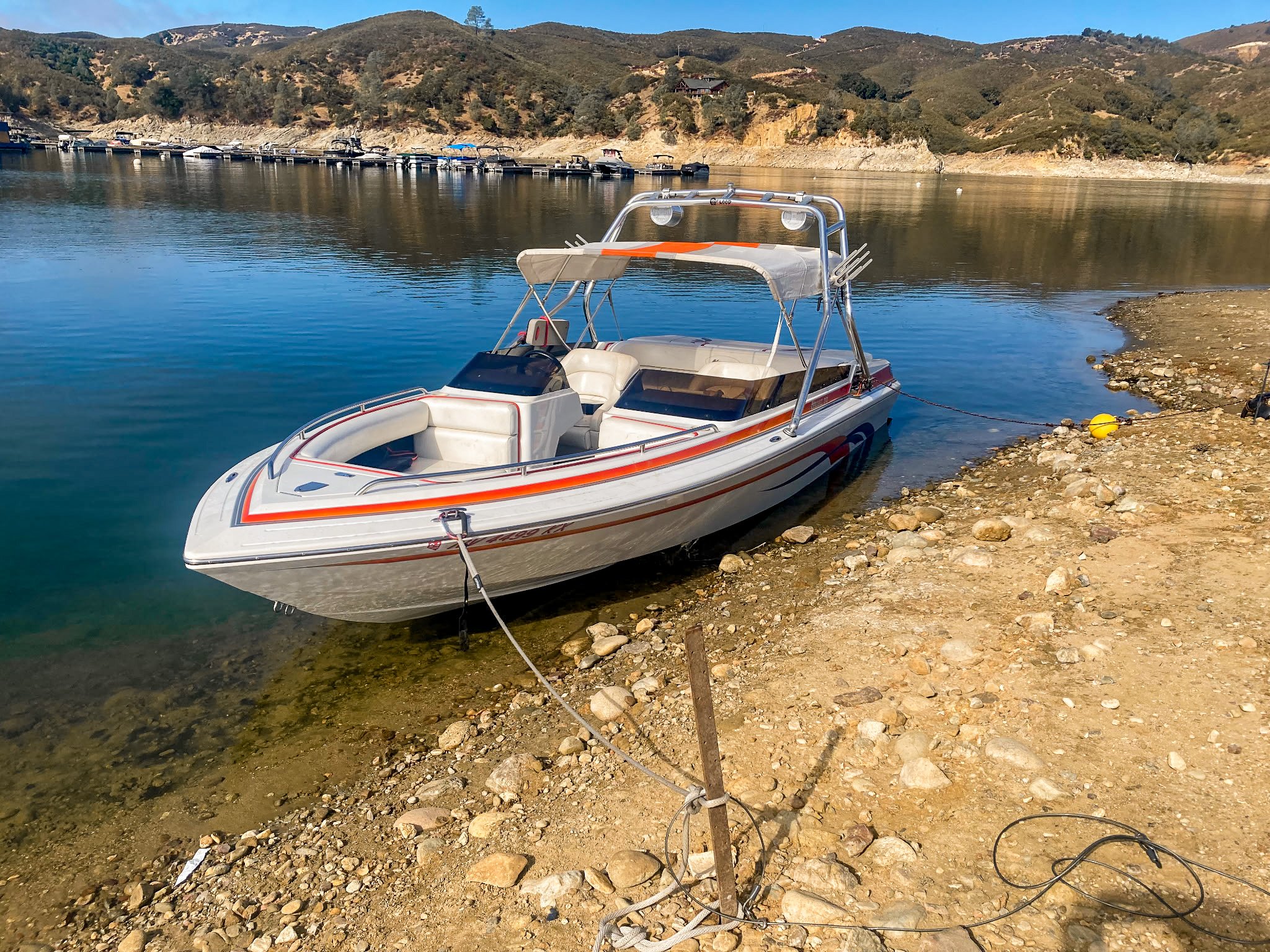 A small motorboat with white seats and an orange stripe is docked near a rocky shore on a calm body of water, with hills and more boats visible in the background.