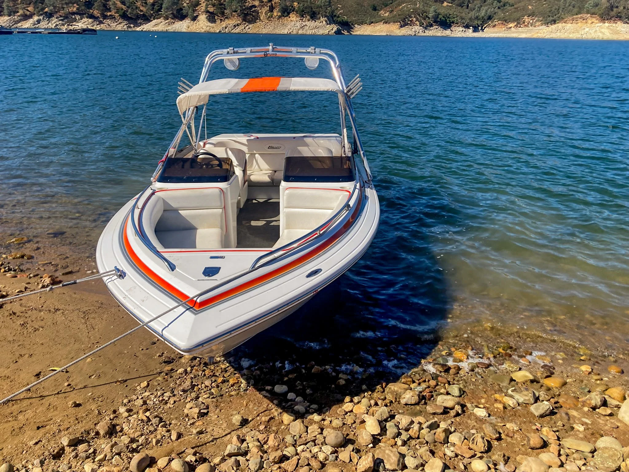 White motorboat with orange and gray stripes is docked on a rocky shore by a lake, with trees and hills in the background.