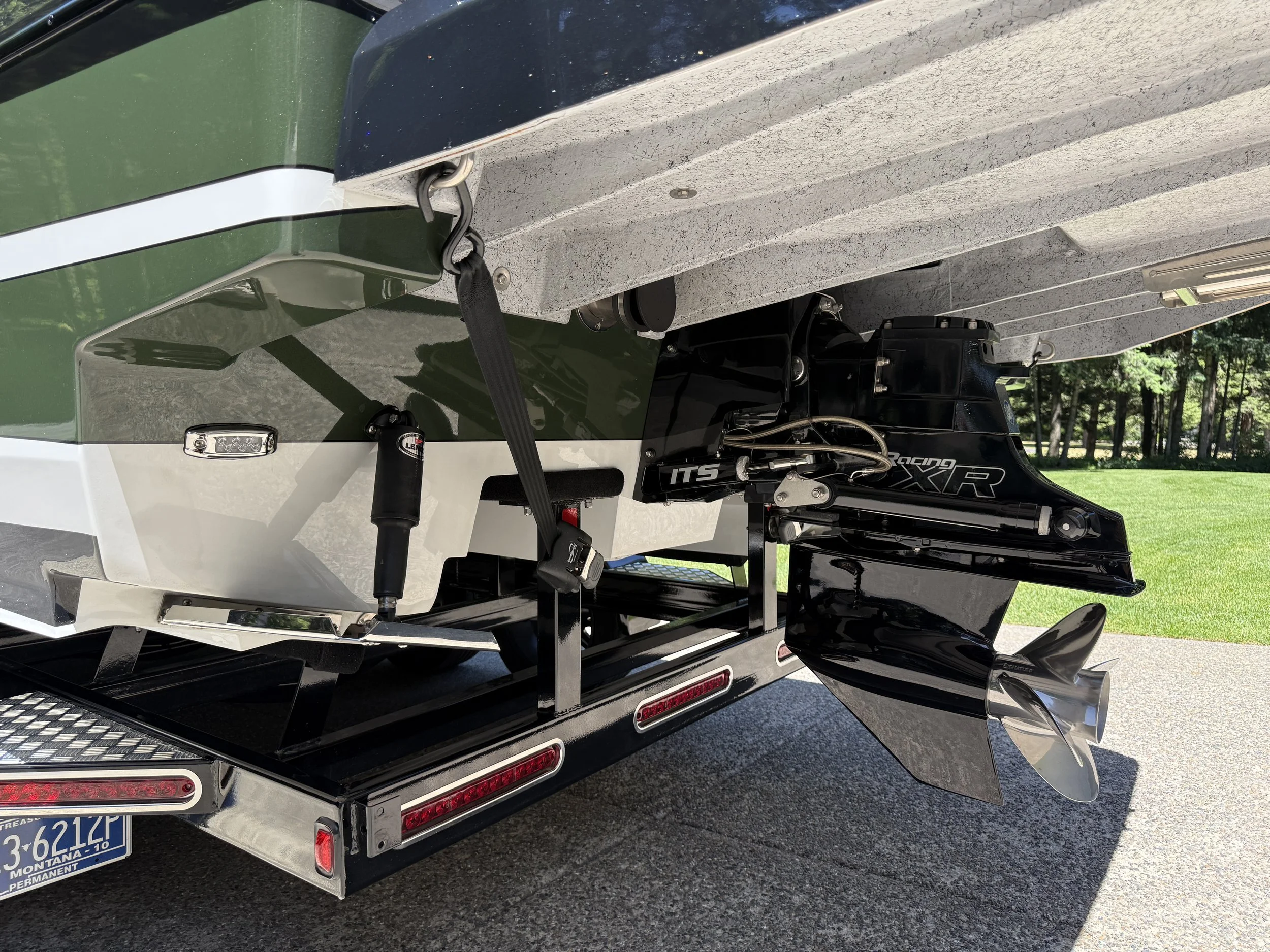 Close-up of the back of a boat trailer showing a boat motor attached to the boat. The boat features a black and green color scheme, a white stripe, and a model number. The trailer has red and white reflective lights and a license plate, with a backgr
