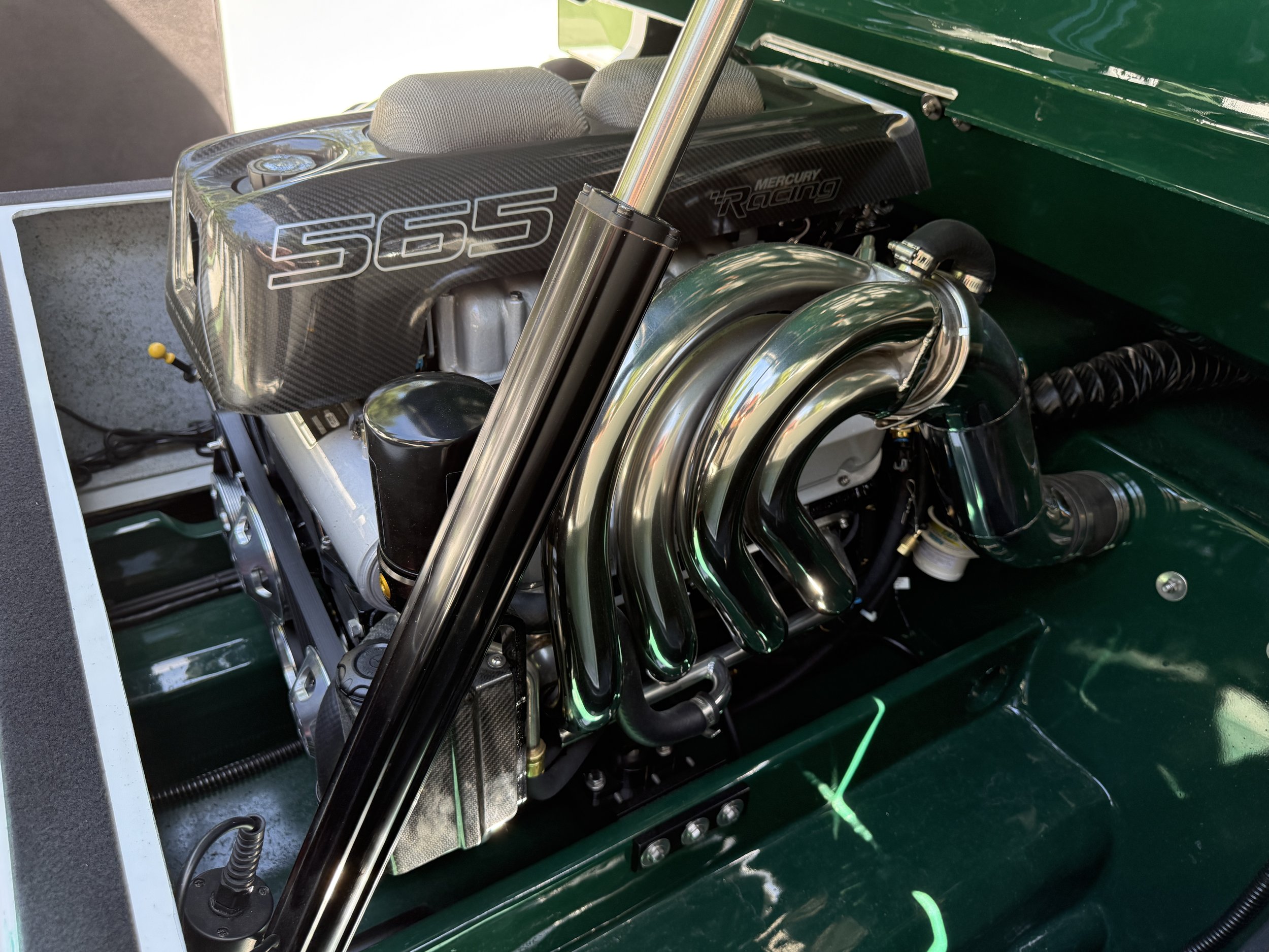 Close-up image of a racing car engine, featuring a carbon fiber cover with the number '565' printed on it, combined with metallic and black components, including a visible turbocharger and suspension parts.