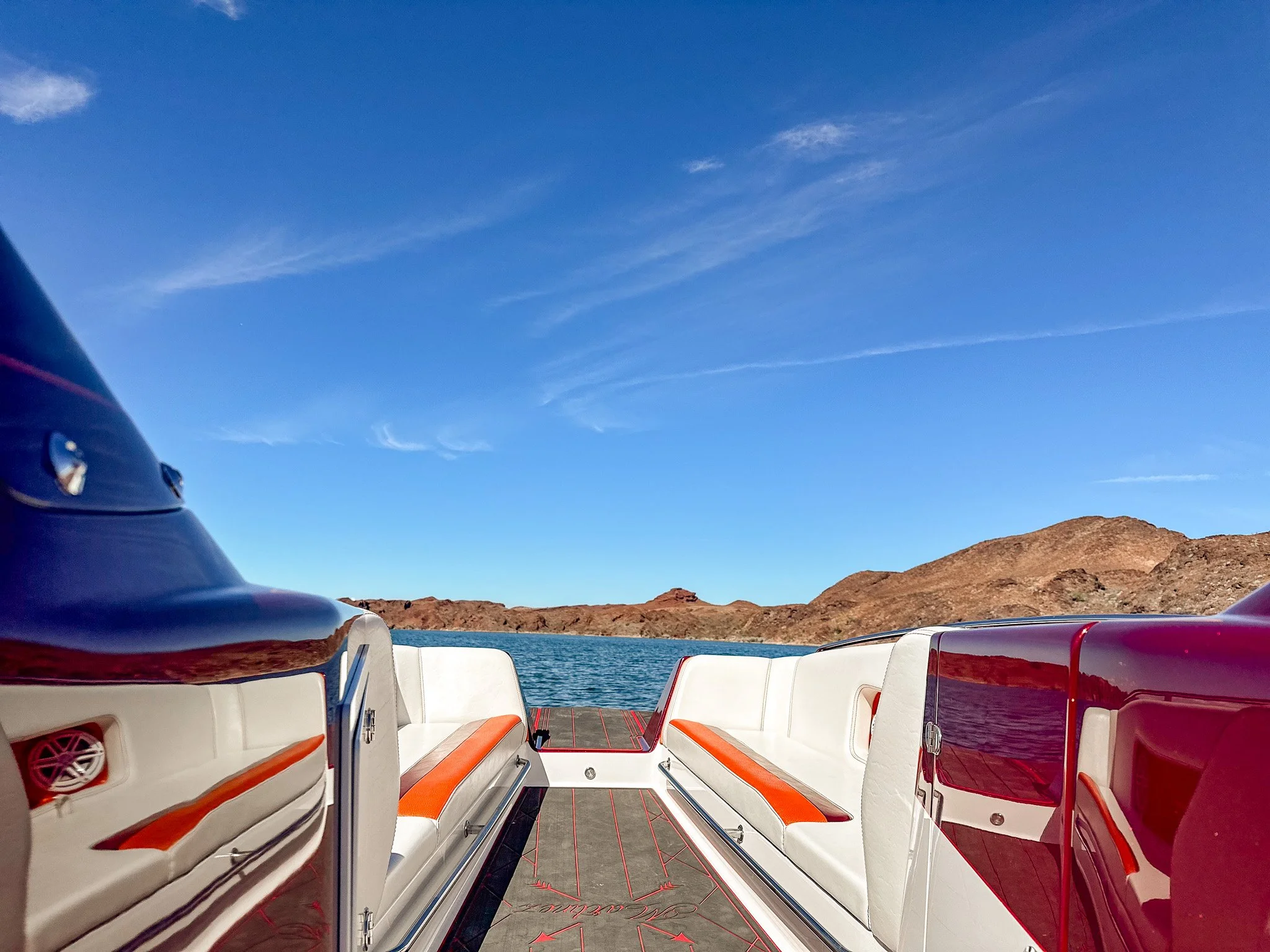 View from the back of a boat showing white and red seating, water, rocky hills, and a clear blue sky with some wispy clouds.