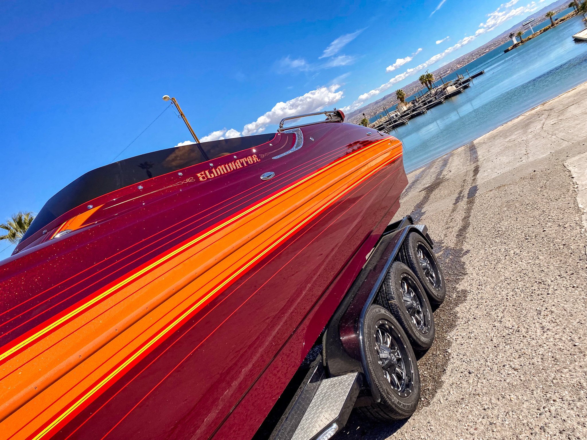 A red boat with orange and yellow stripes on the side, docked on a sandy shore near a body of water with multiple docked boats and palm trees in the background, under a partly cloudy sky.