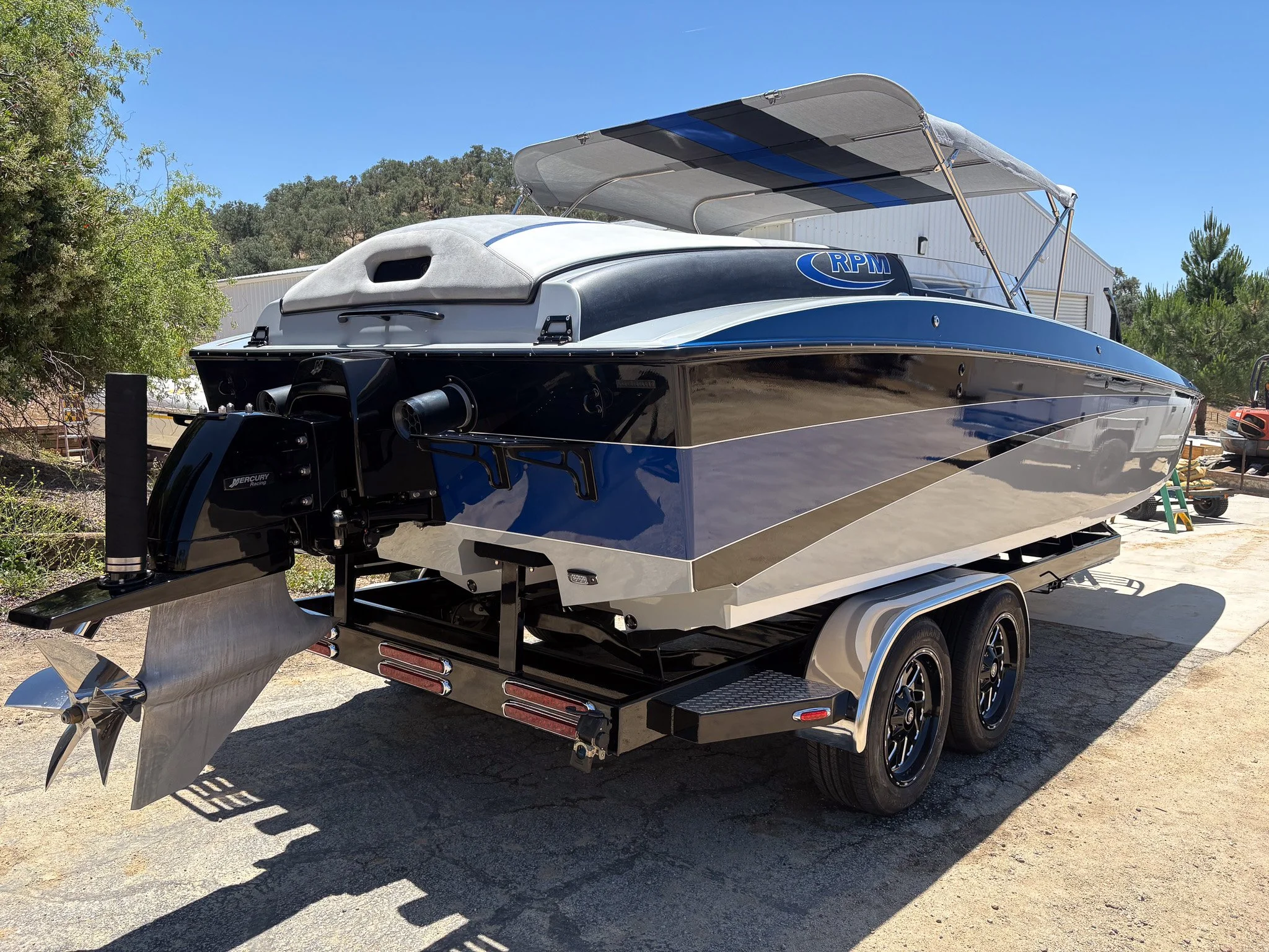 A sleek blue and white speedboat on a trailer, with an outboard motor and a canopy, parked outdoors near trees and a building under a clear blue sky.
