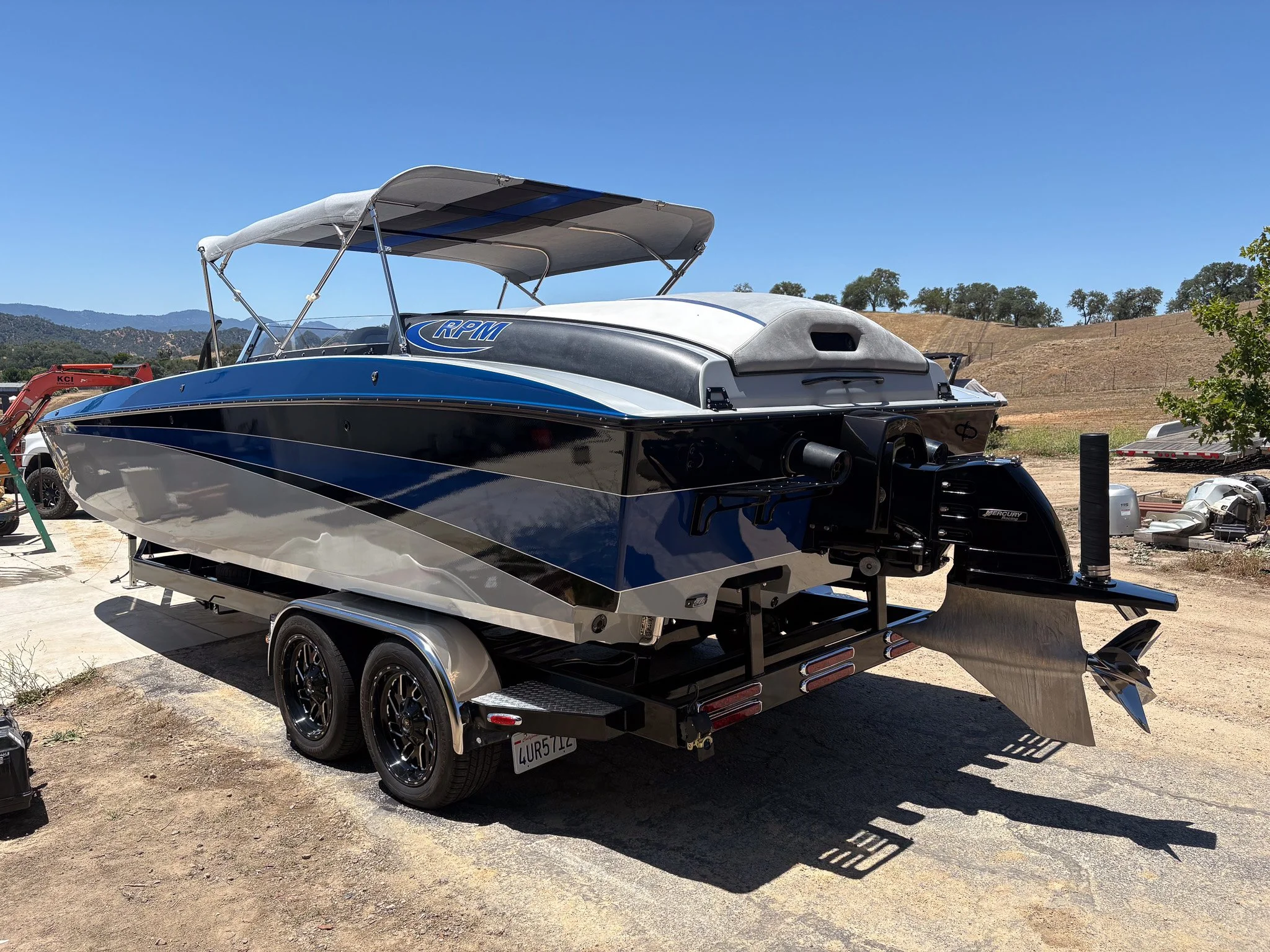 A black and blue speedboat on a trailer in an outdoor area with dirt ground, surrounded by trees and hills in the background, under a clear blue sky.
