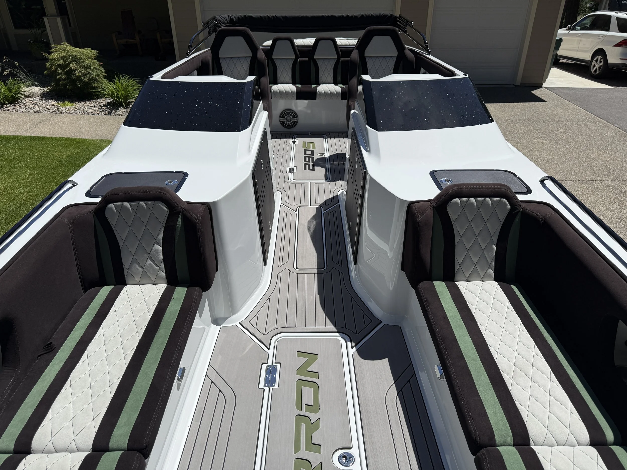 The image shows the interior of a white boat with multiple black and white quilted seats, a gray and green patterned floor, and a black roll cage at the top. The boat is docked outside in a residential area with a garage, a driveway, and a car visibl
