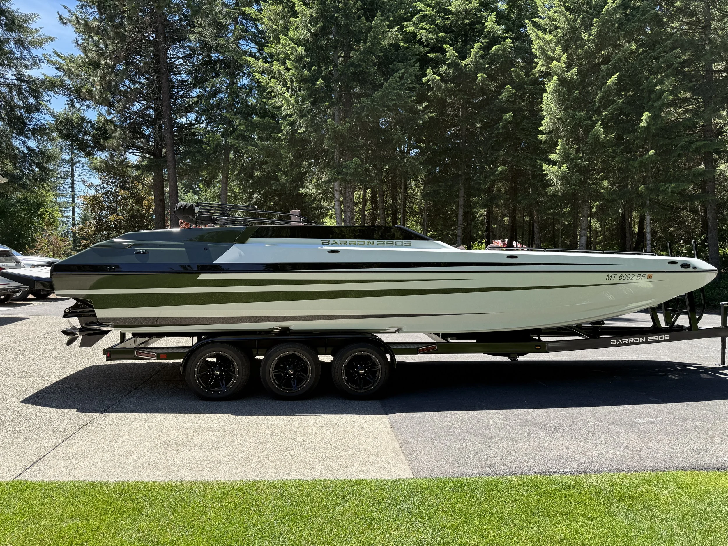 A speedboat on a trailer parked on a concrete driveway, with a background of tall green pine trees and blue sky.
