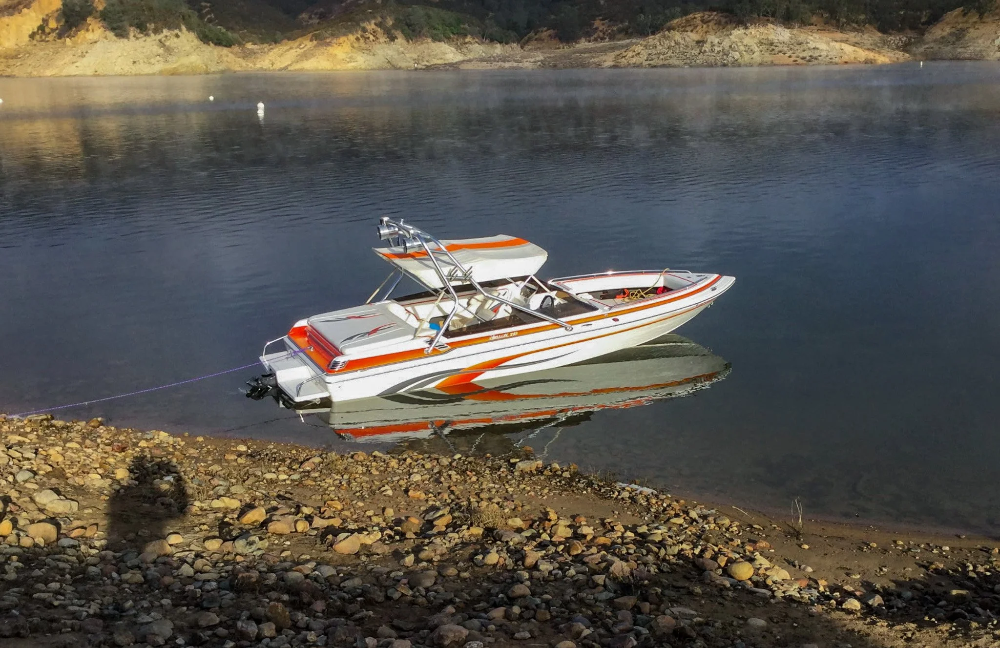 A white and orange speedboat floating on calm water near a rocky shoreline, with a hilly landscape in the background under a mostly clear sky.