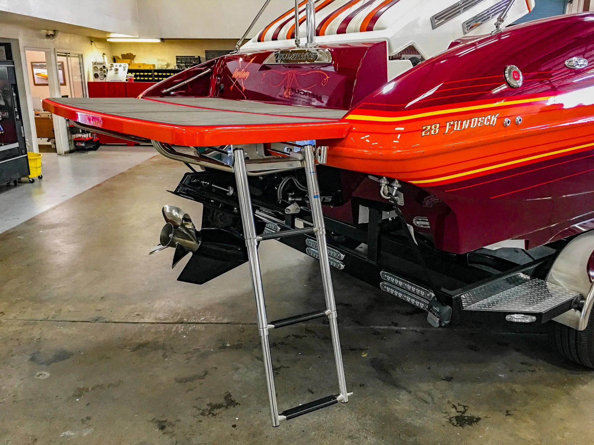 A red race boat with yellow and orange stripes and the number 28 FuDeck written on the side, situated inside a garage or workshop with tools and equipment visible in the background.