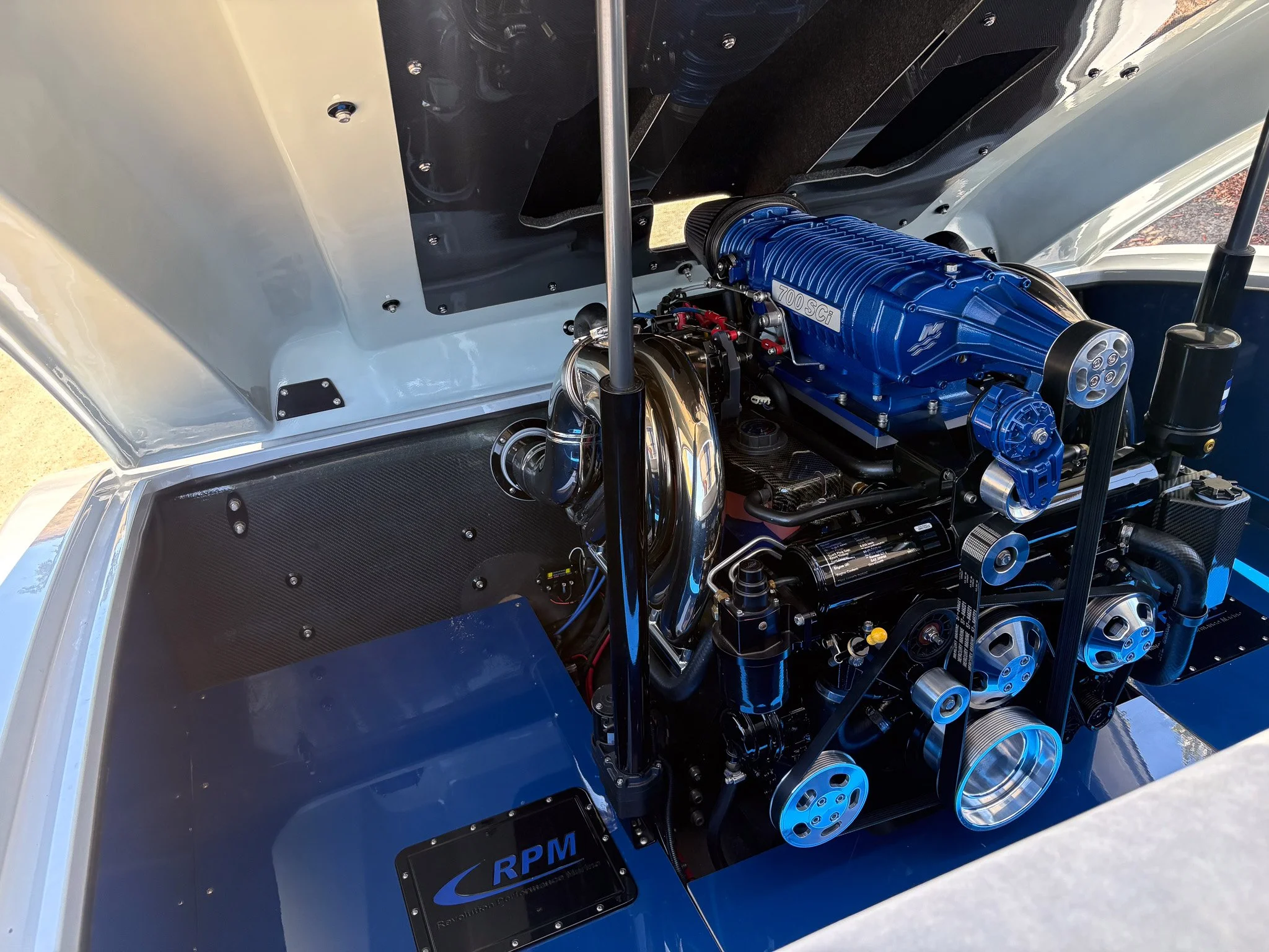 A close-up interior of a race car engine bay, showing a blue supercharger labeled '700 SCI', black and silver mechanical components, pulleys, and belts.