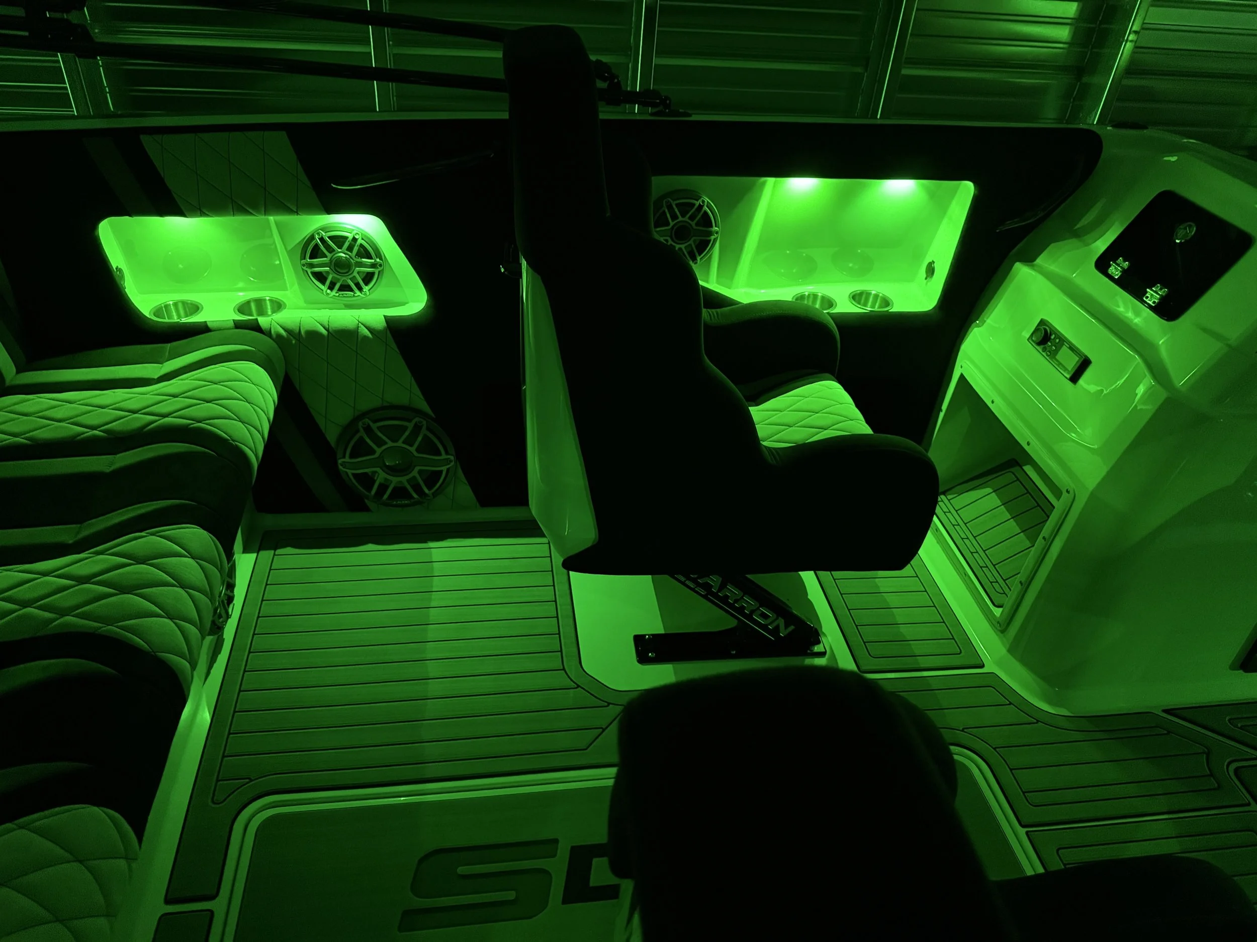 Interior of a boat with black seating, illuminated by green LED lighting, speaker system, control panel, and wood-like flooring.