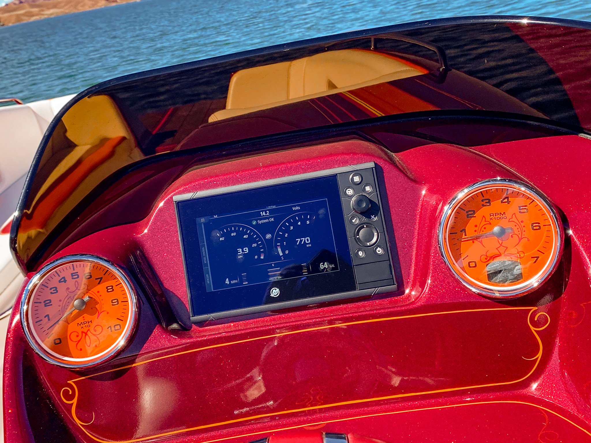 The image shows the dashboard of a boat with two bright orange gauges, a digital display, and a deep red console with black and gold accents. The boat is on a body of water, with a shoreline visible in the background.