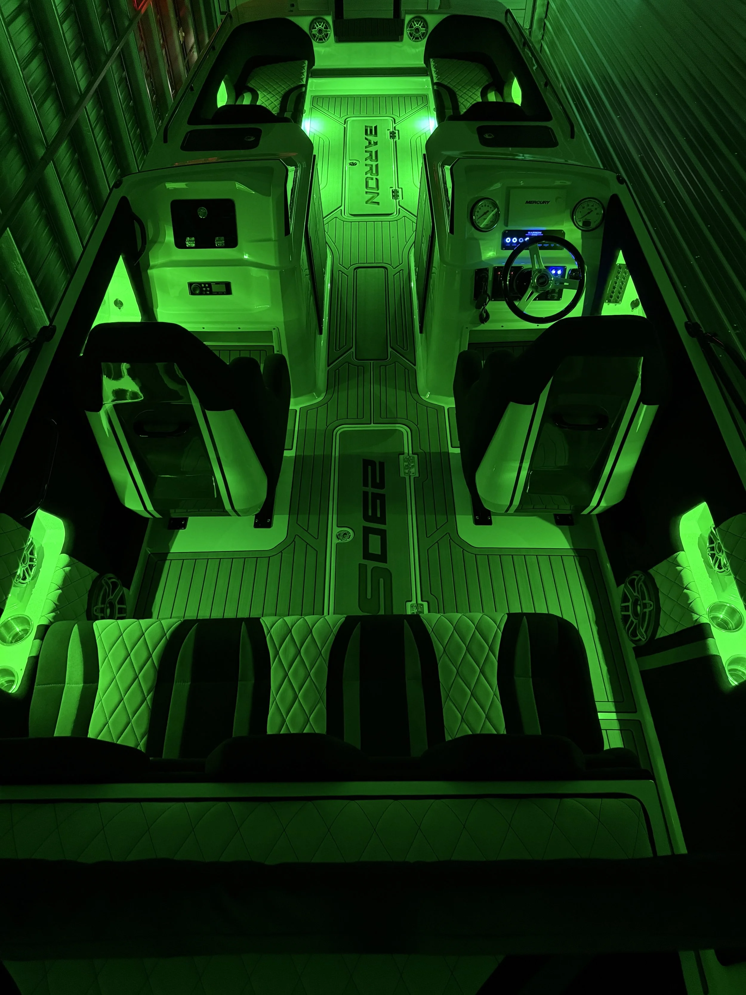A boat interior illuminated with green LED lights, featuring black and white quilted seats, a steering wheel, gauges, and a central storage compartment with the word 'BARRON' on it.